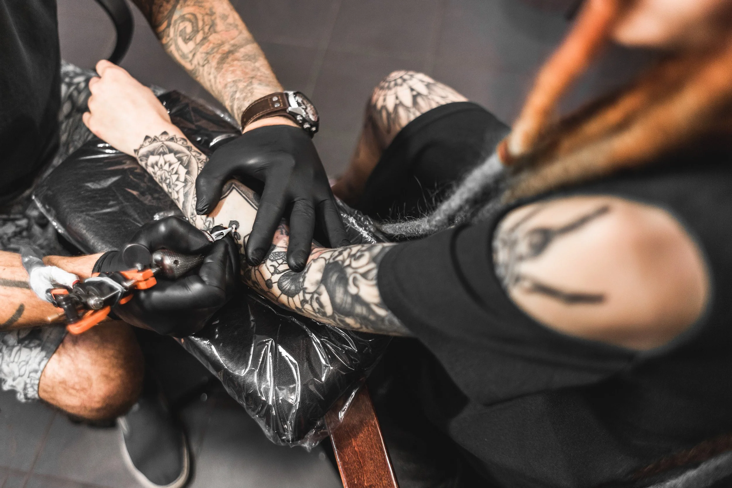 A tattooed person getting a tattoo on their arm by a tattoo artist wearing black gloves.