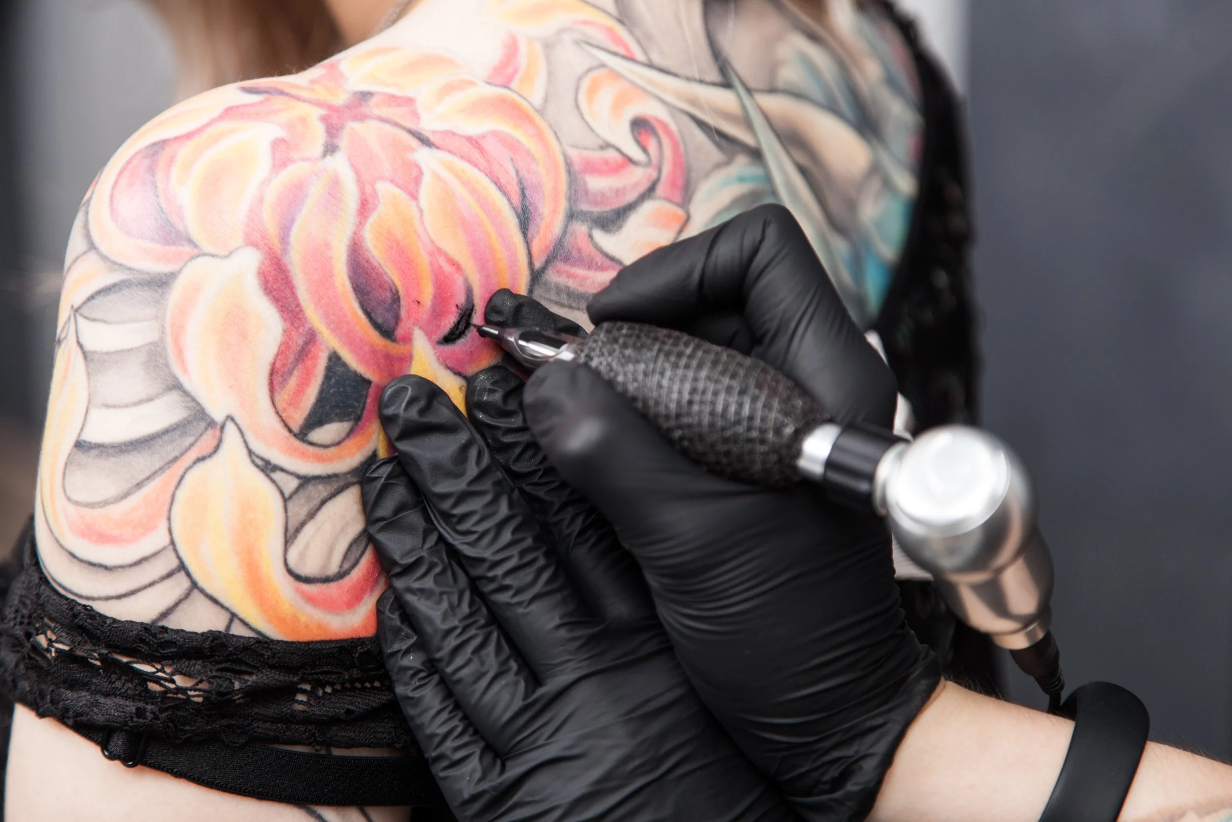 A person with colorful floral tattoos on their shoulder is getting a tattoo done by an artist wearing black gloves, using a tattoo machine.