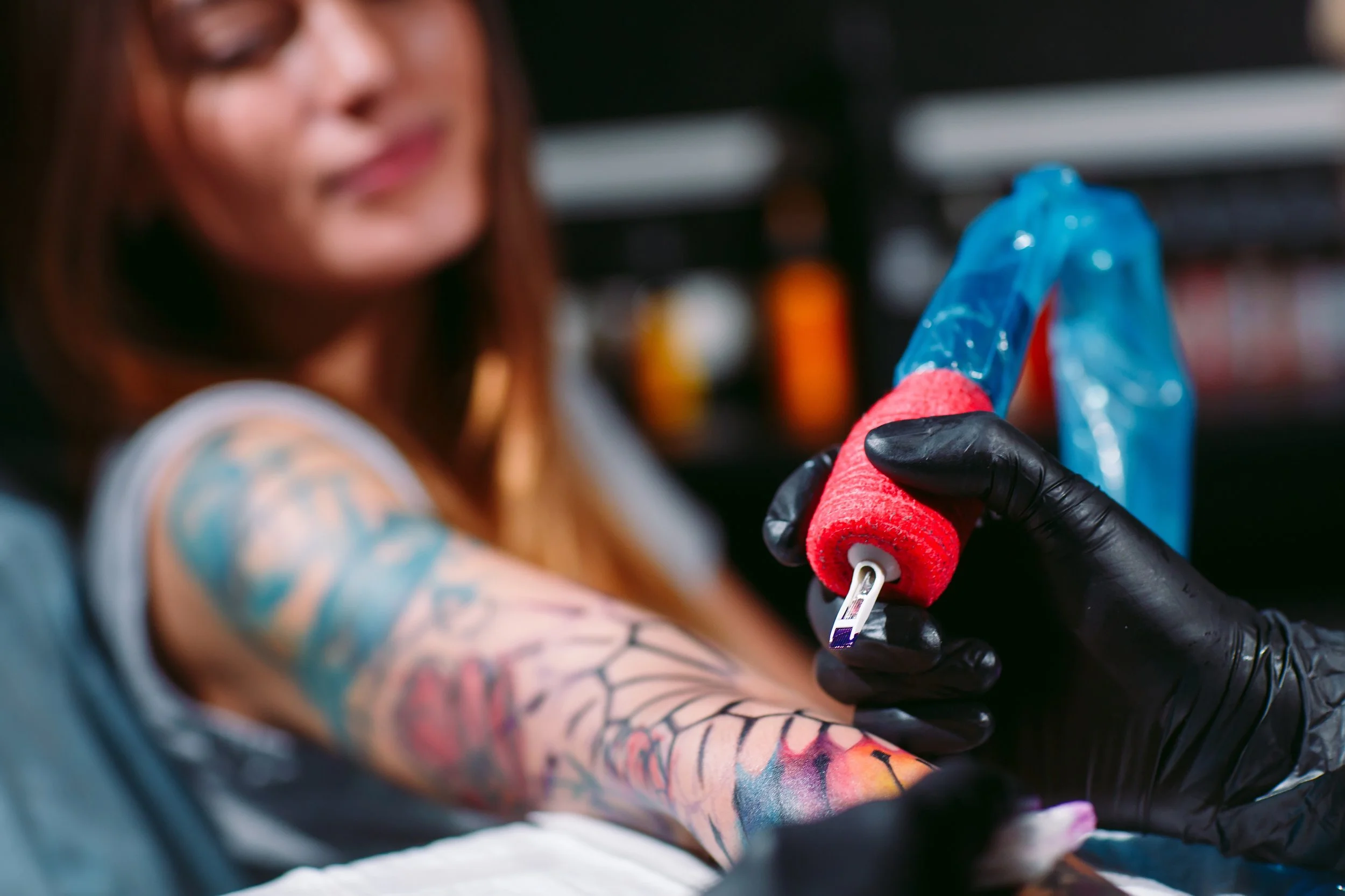 Tattoo artist using a machine with a red grip to tattoo a person's arm, who has colorful tattoos, in a tattoo studio.