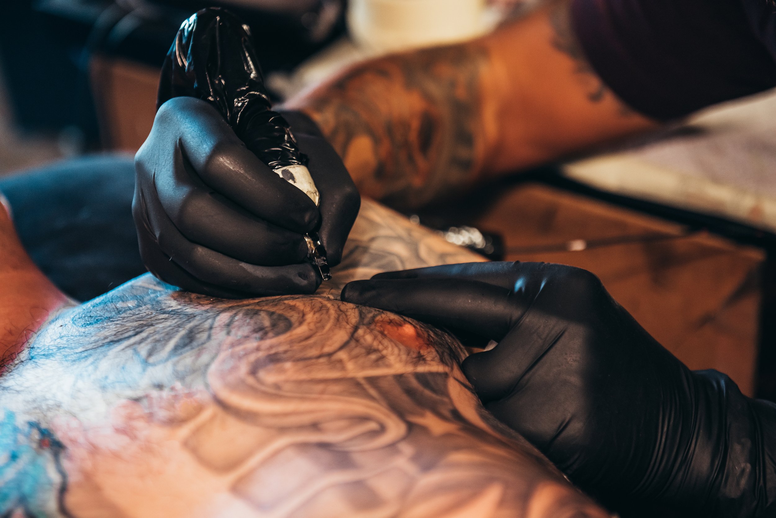 A tattoo artist in black gloves is creating a colorful tattoo on a person's arm with visible tattoos and inked skin.