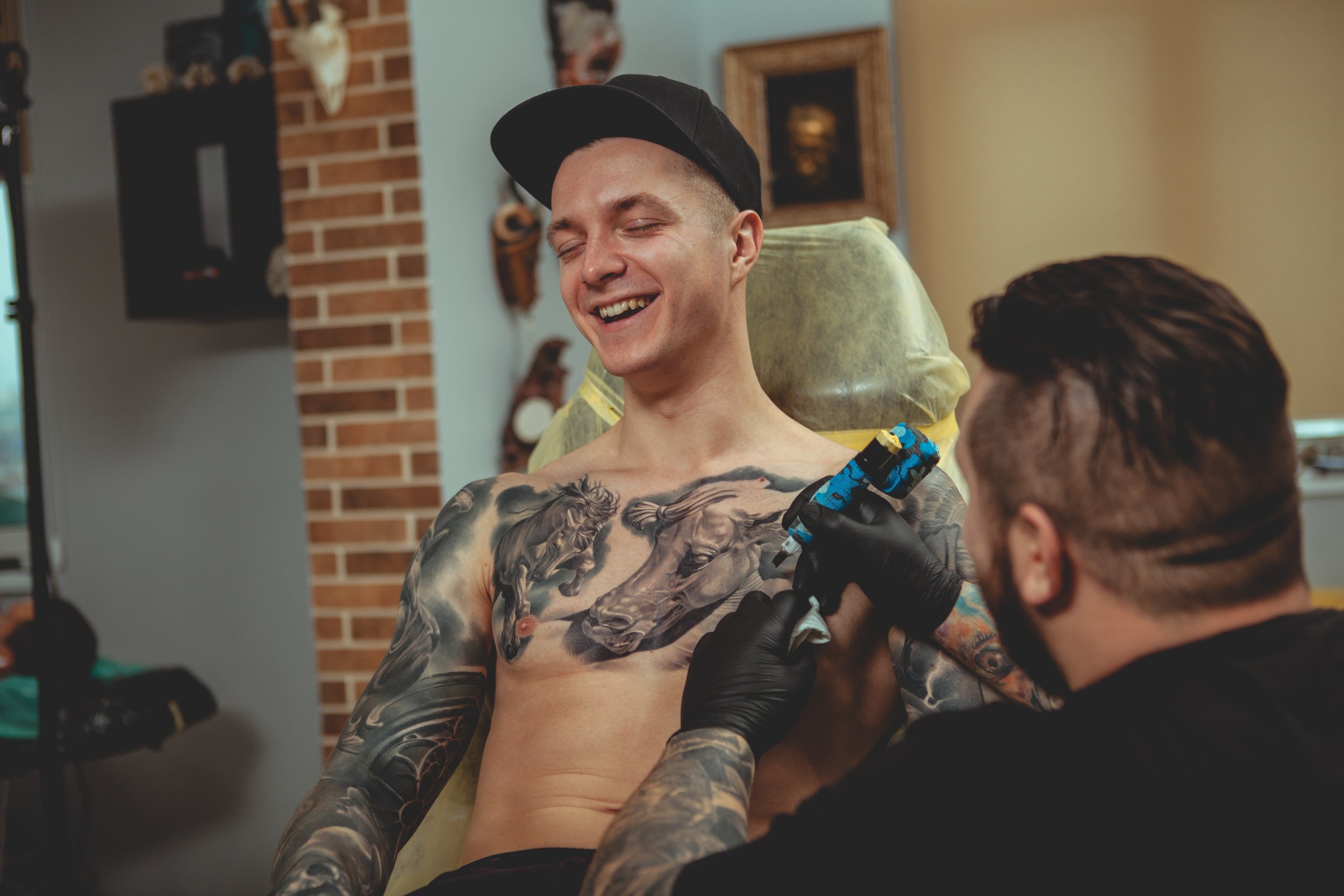 A man with tattoos on his chest and arms is smiling while getting a tattoo of a lion's head on his chest from an artist in black gloves. The man is wearing a black cap and sitting on a chair covered with yellow protective paper.