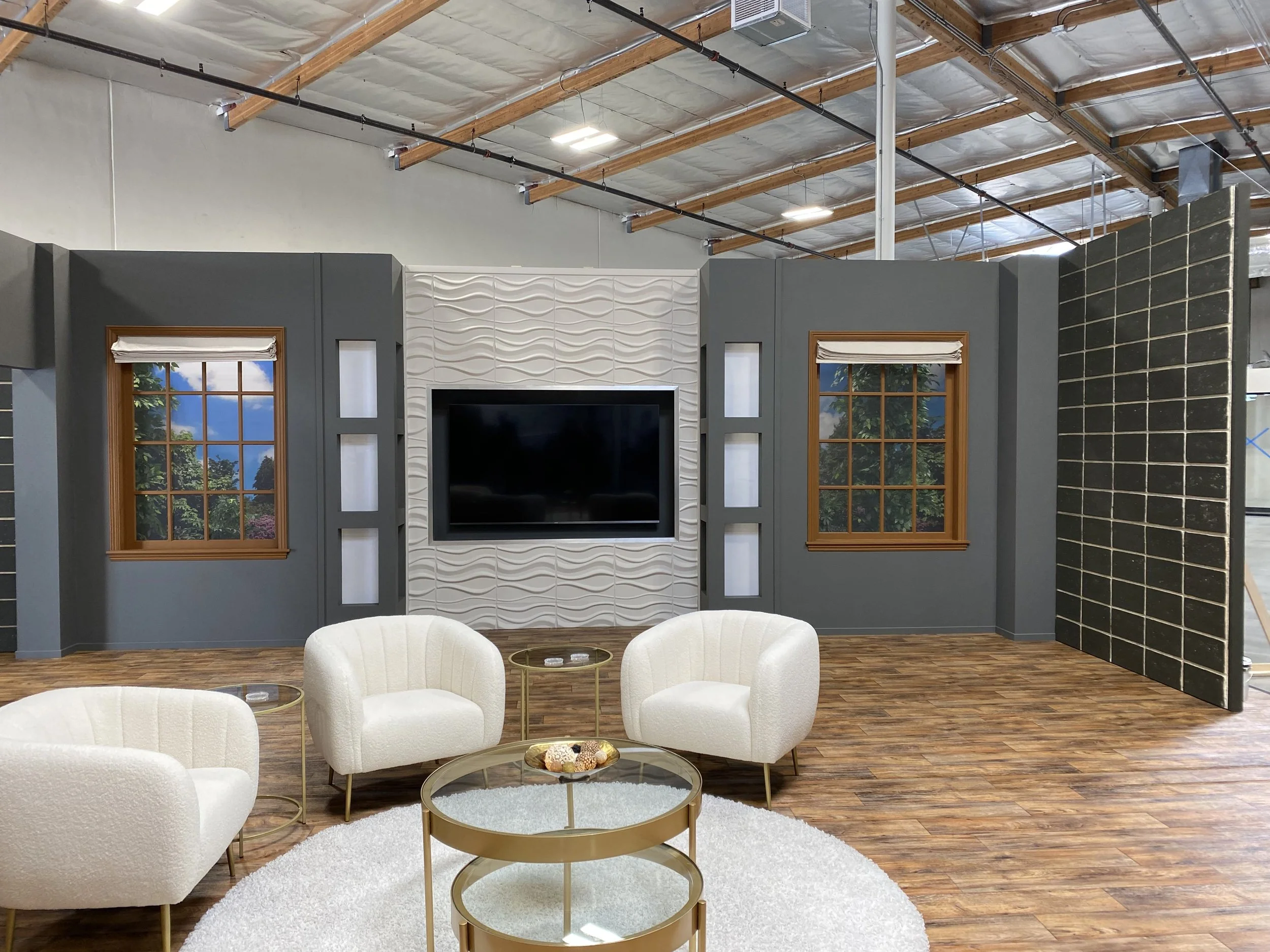 Indoor studio setup with a gray wall backdrop, two windows with wooden frames showing outdoors, a mounted flat-screen TV, and white seating with a round gold coffee table in front.