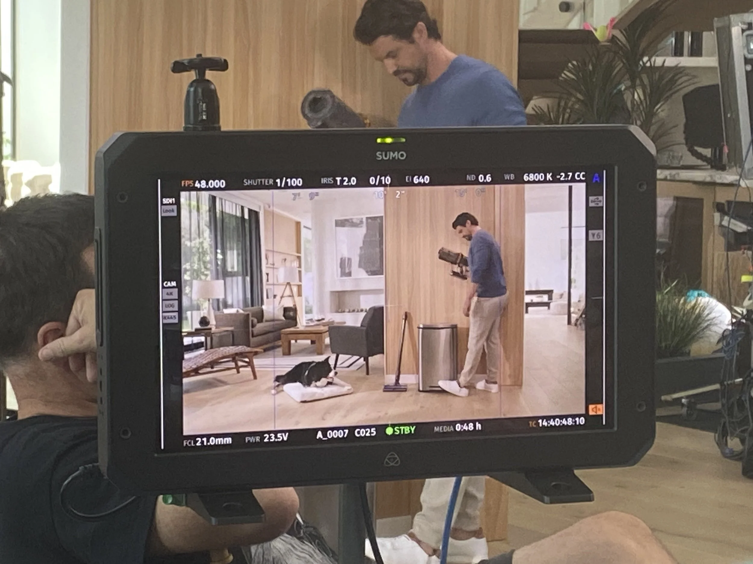 Monitor on set showing a scene of a man holding a video camera, inside a modern apartment with wooden accents, large windows, and a black and white dog lying on a cushion.