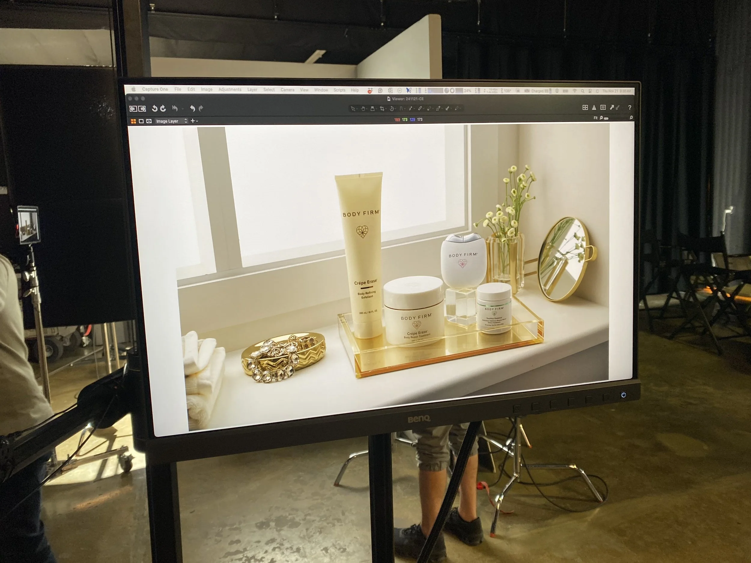 Close-up of a tabletop displaying skincare and beauty products, including a tube labeled 'BODY FIRM,' a jar, a candle, and a small mirror, set in front of a window with natural light.