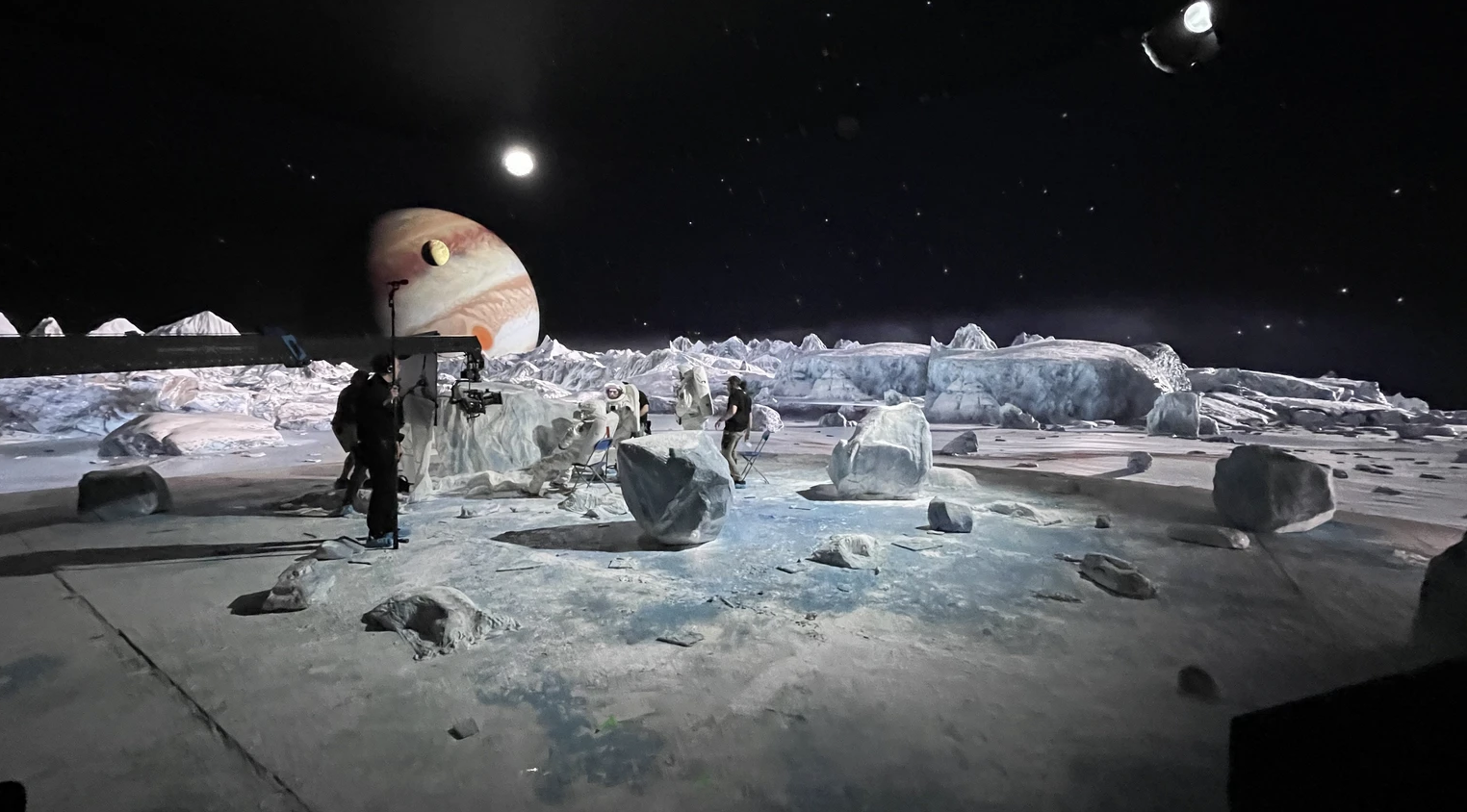A film or photo shoot set designed to look like an icy, extraterrestrial landscape with actors and crew members, ice formations, and planets in the background.