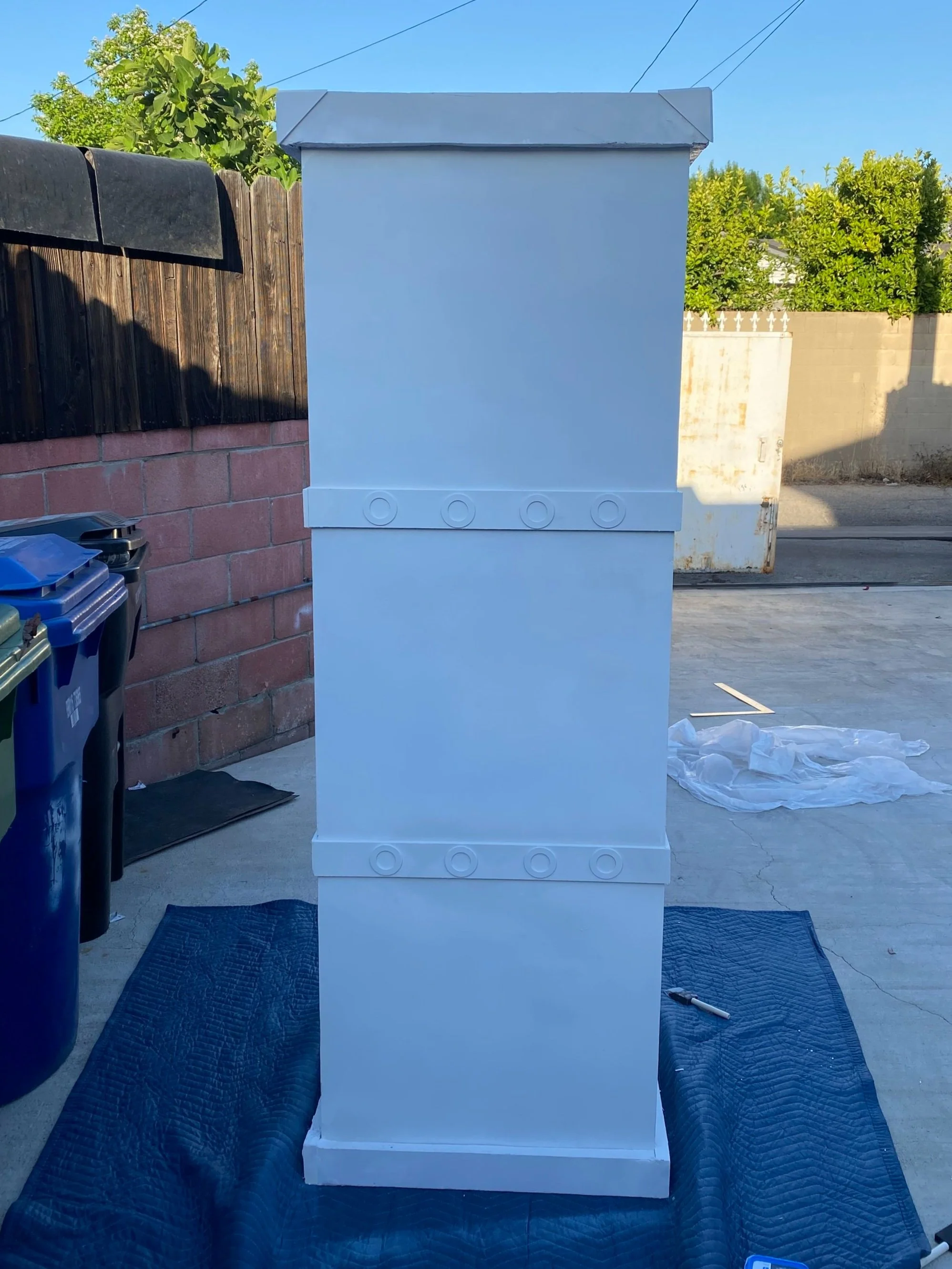 White, painted, rectangular outdoor cabinet with circular details on two horizontal bands, set on a dark blue blanket on concrete ground in a backyard, with trash bins, a brick wall, and green trees in the background.