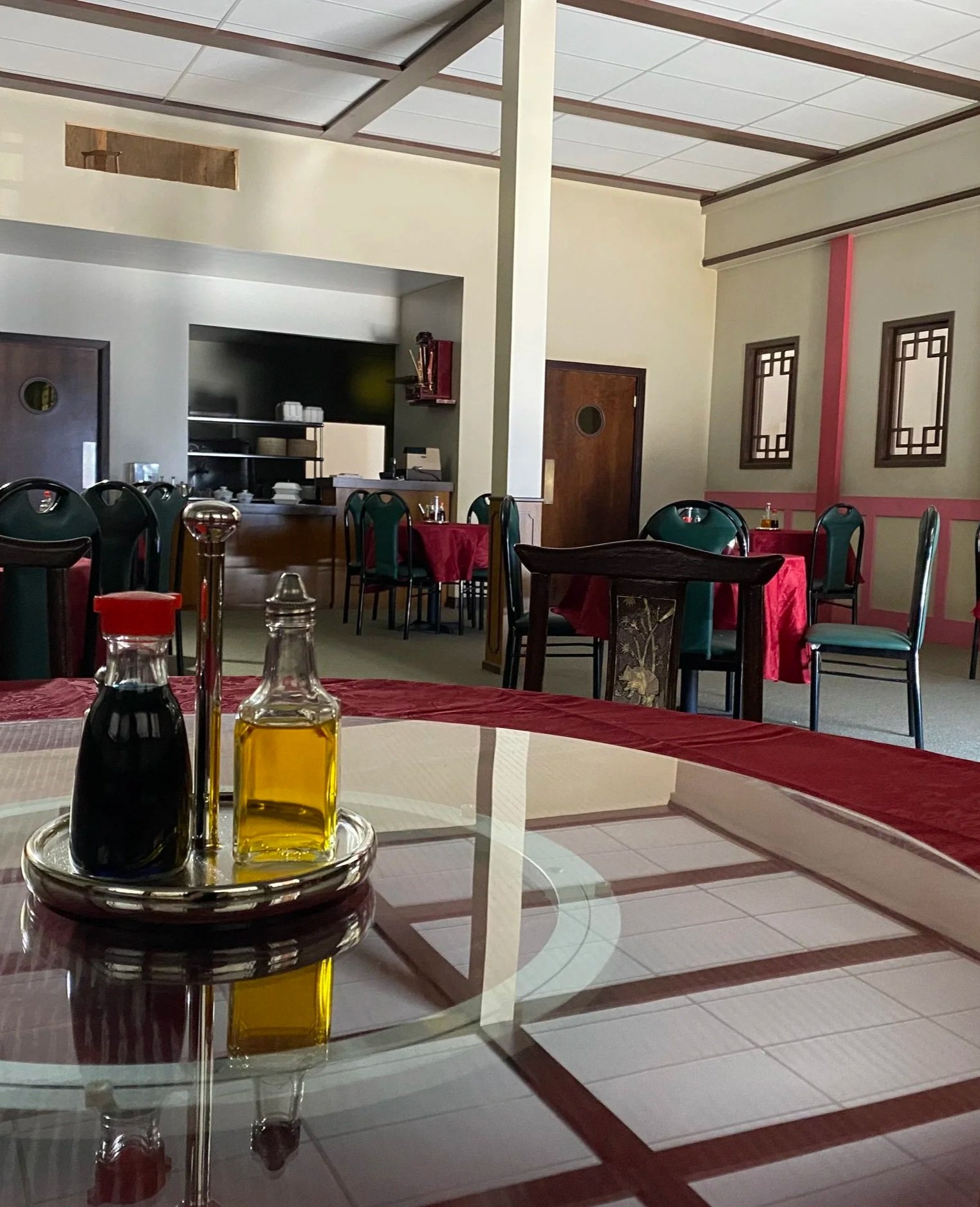 Restaurant dining area with tables covered in red tablecloths, green chairs, and a condiment set with soy sauce and oil on a glass table in the foreground.
