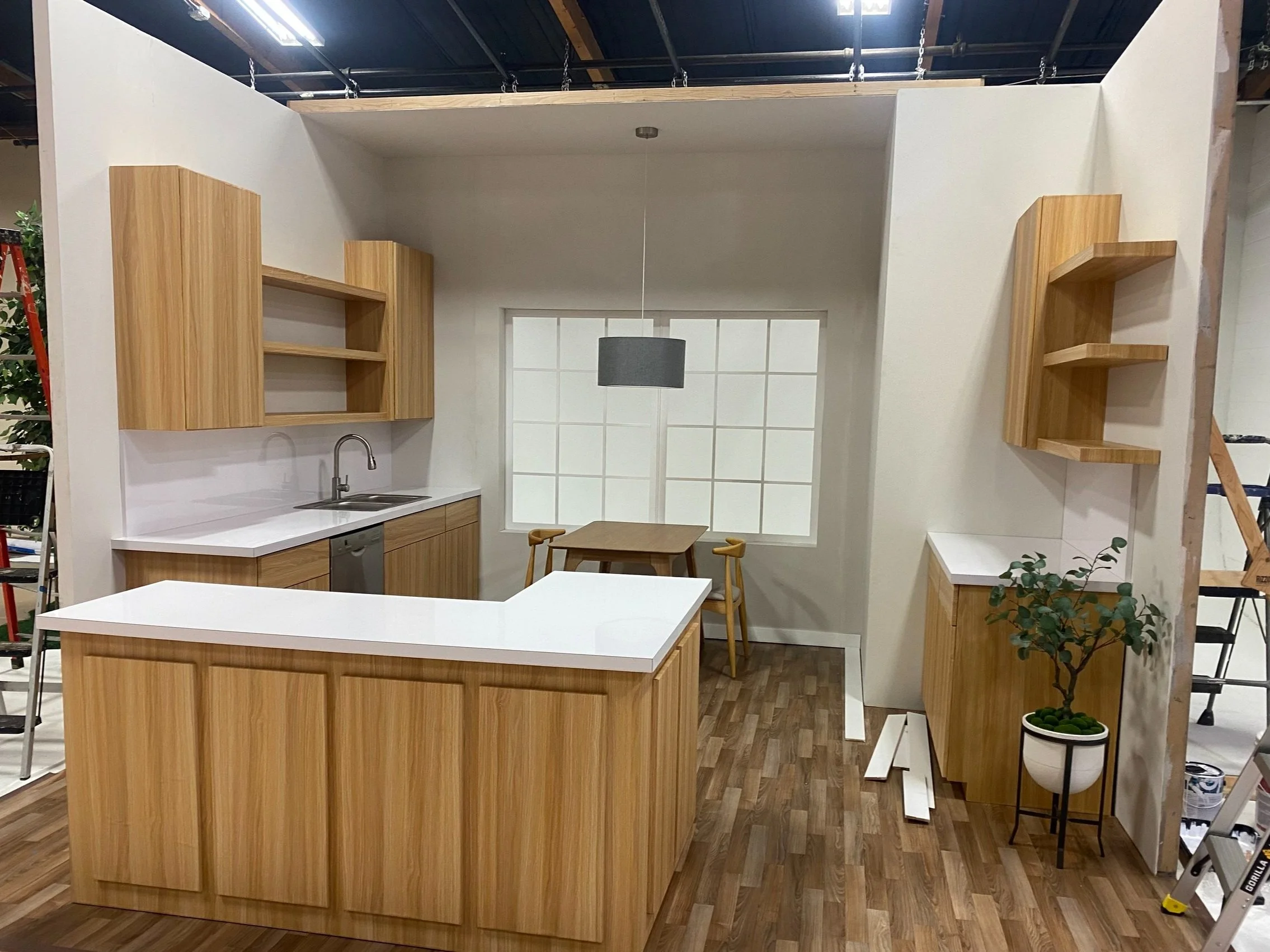 A modern kitchen display with wooden cabinets and white countertops, a small dining table with two chairs, and a potted plant, all set in a room under construction with ladders and building materials present.