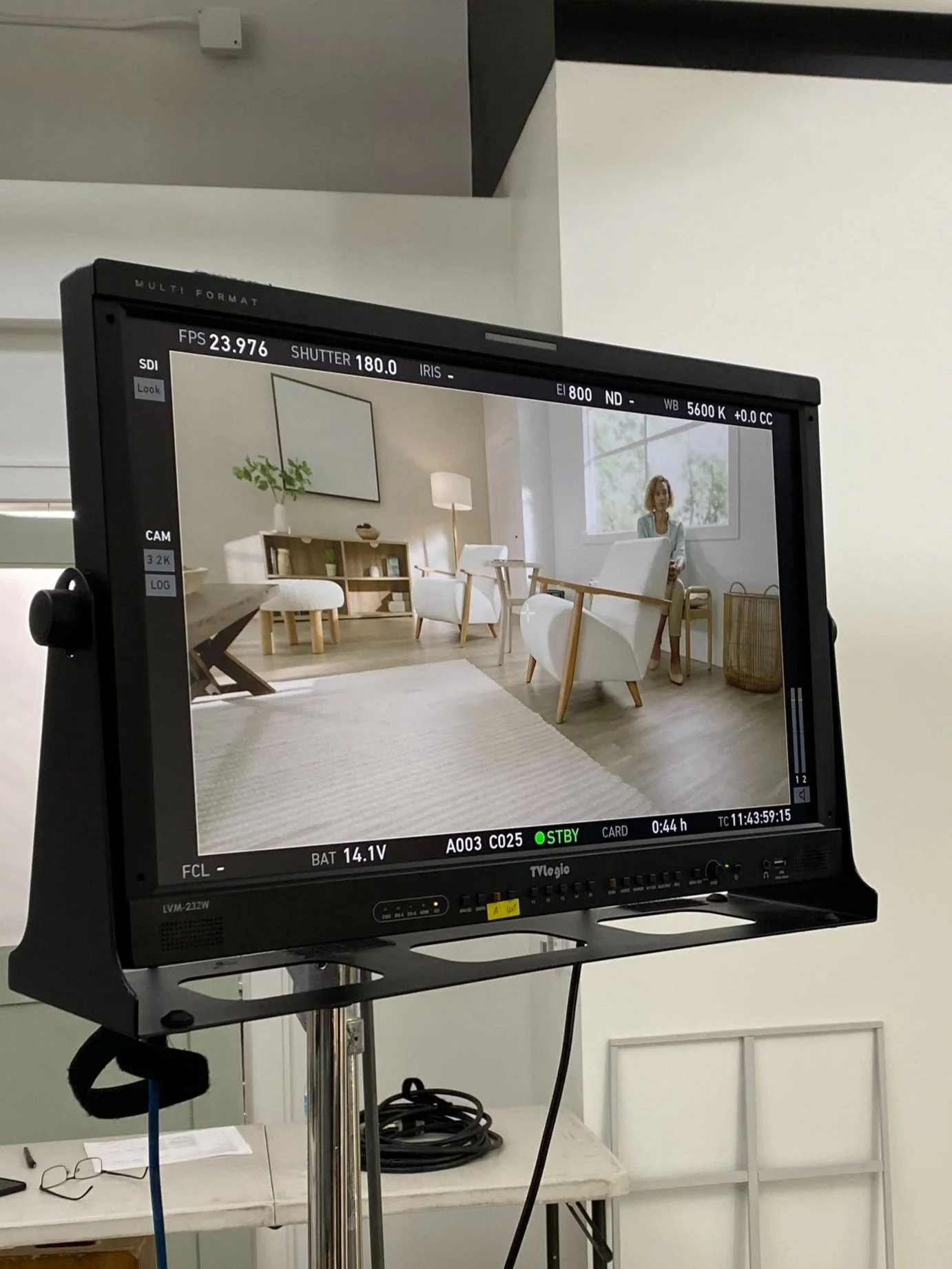 A professional video camera monitor displaying a shot of a bright living room with modern furniture, including chairs, a table, a plant, a lamp, and a woman sitting near a window.
