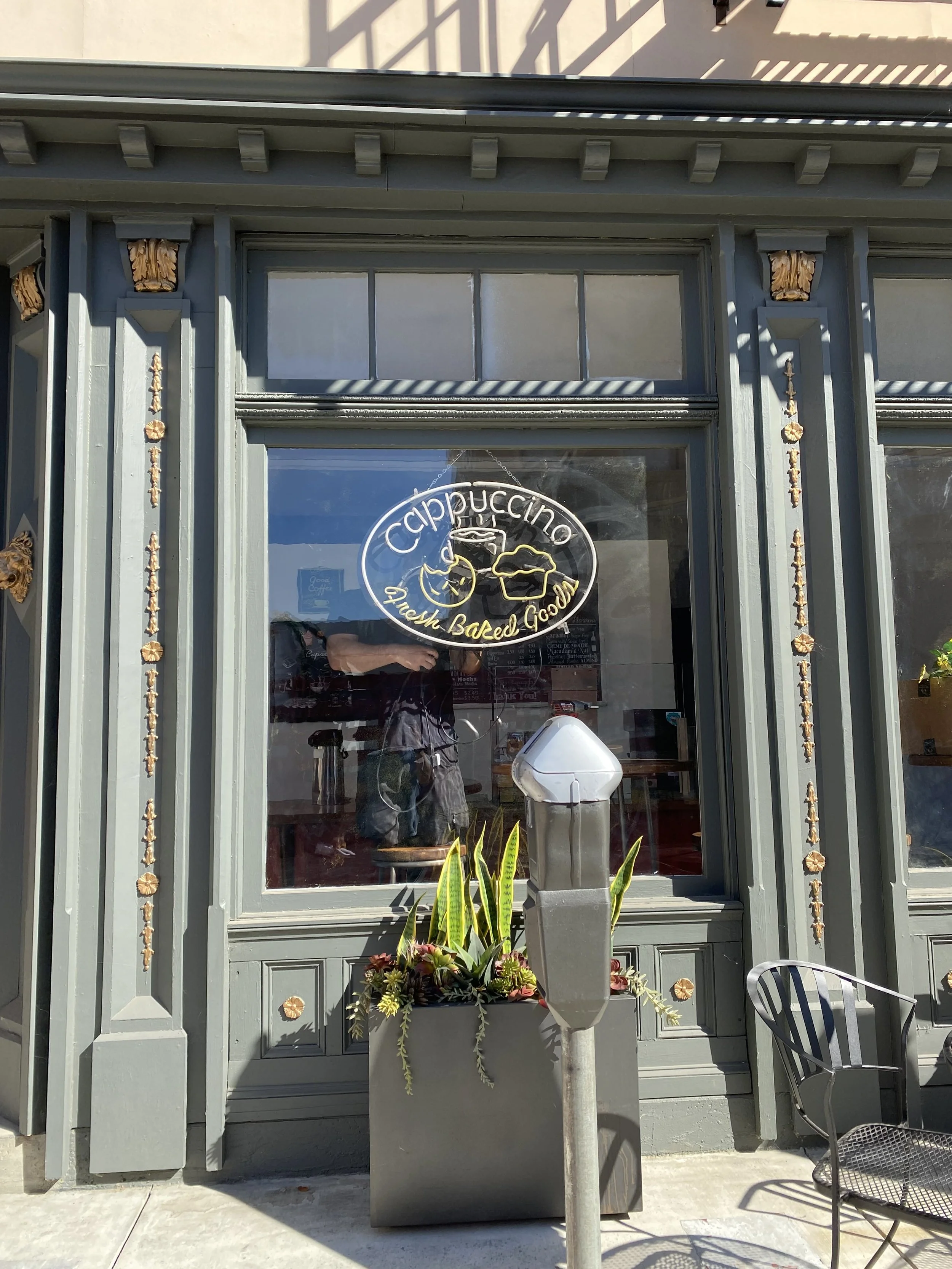 Exterior of a green-gray storefront with a circular neon sign reading 'Cappuccino' and 'Fresh Baked Goods', a large planter with green plants, a black metal chair, and a parking meter in front.