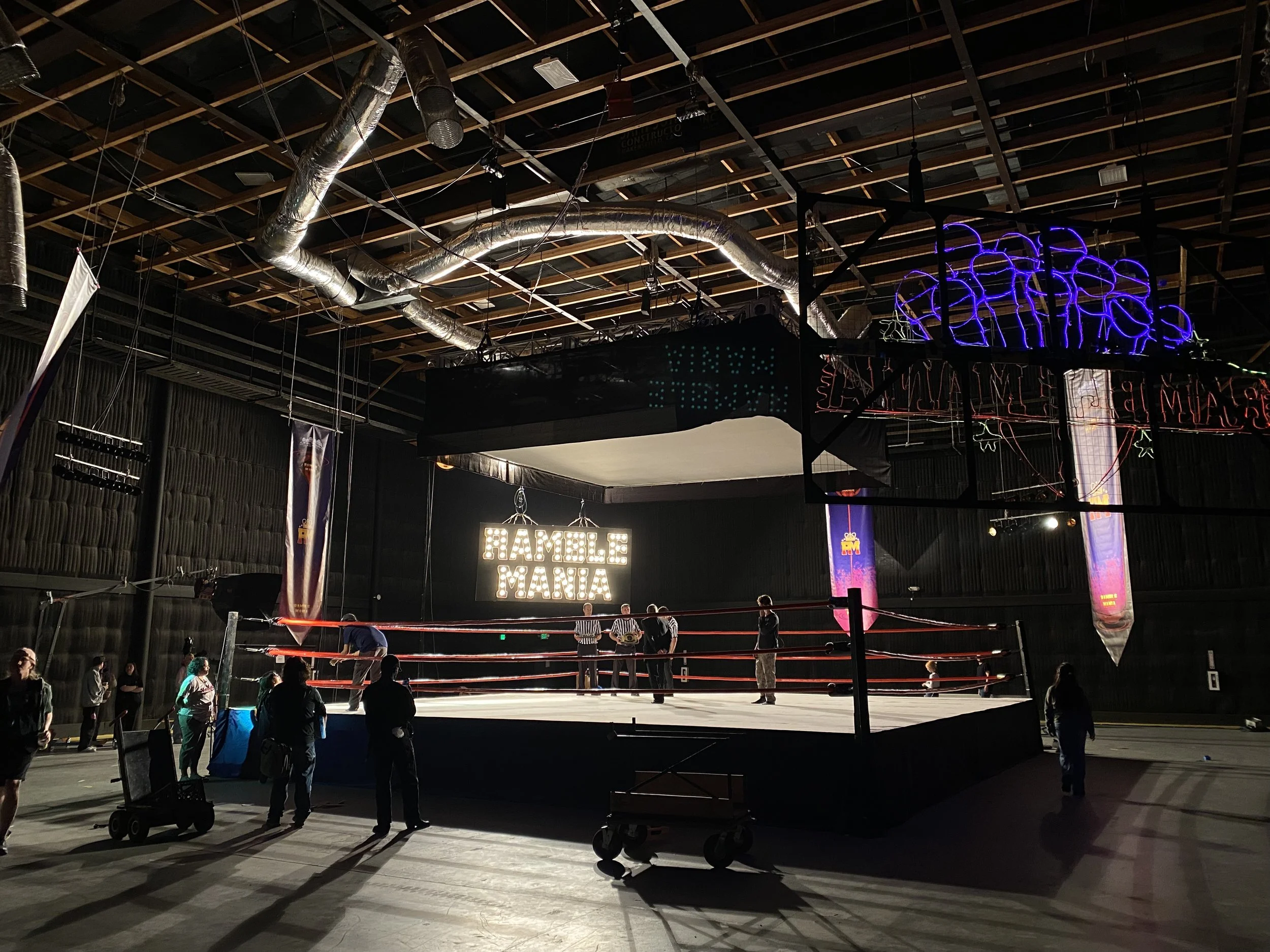 Indoor professional wrestling ring with several people around it, illuminated by marquee sign reading 'Hamble Mania' in the background, with dim overhead lighting.