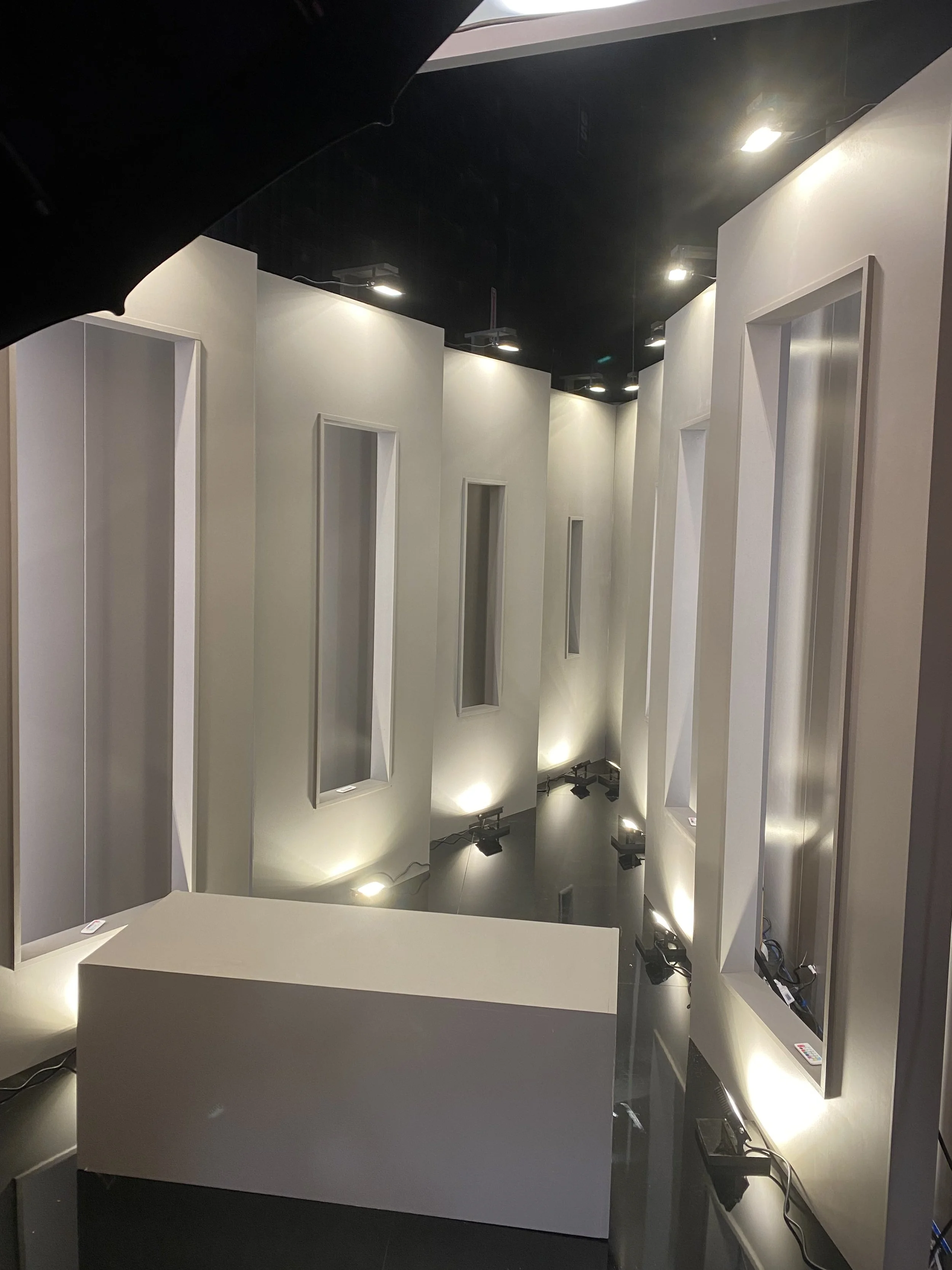 Interior of a modern room with tall vertical white panels with inset rectangular displays, illuminated by small spotlights, and a white desk at the front.