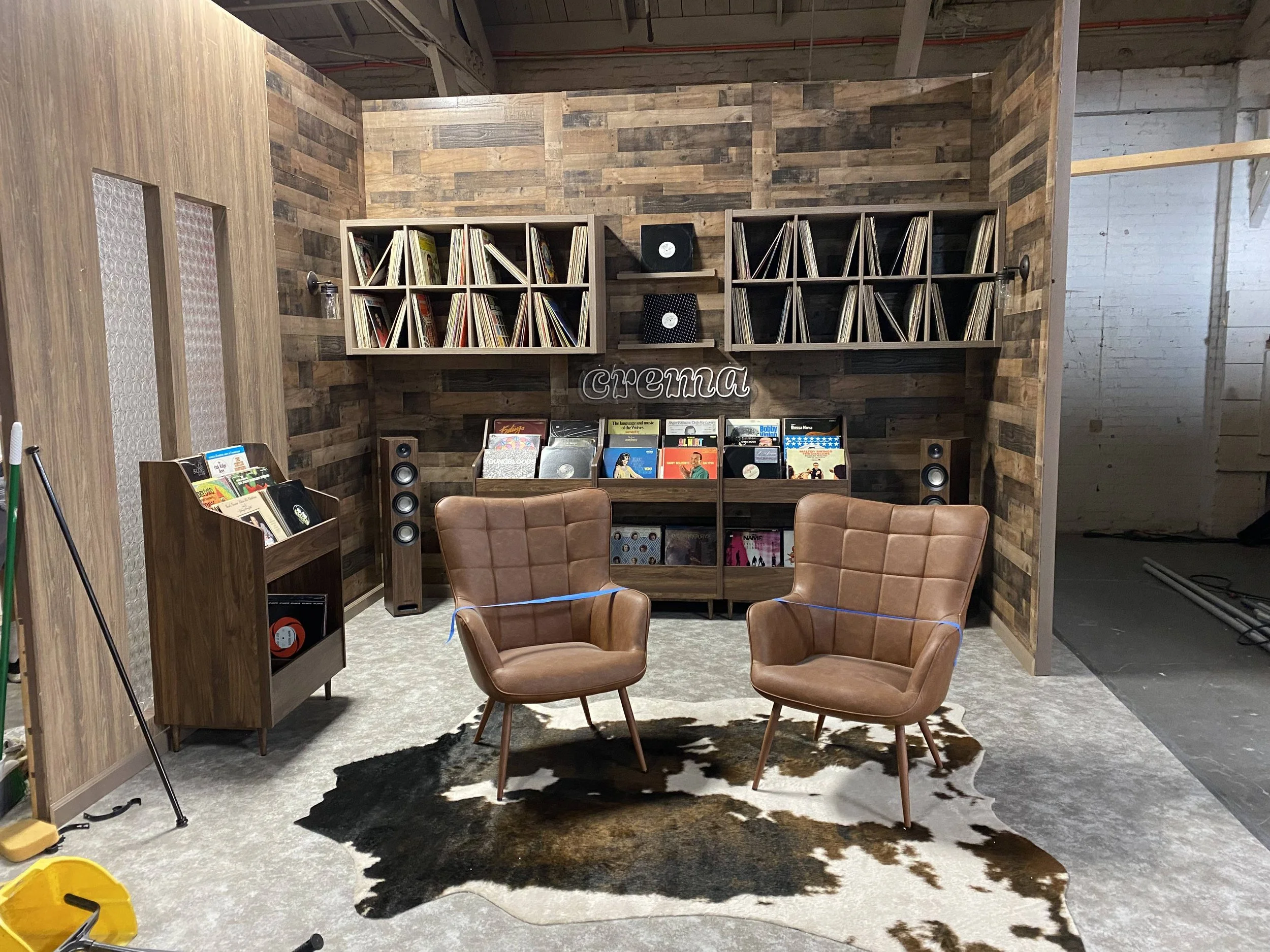 A cozy room with a vintage record collection, two brown leather armchairs, a cowhide rug, and wooden wall paneling, with a 'cinema' sign and stereo equipment.