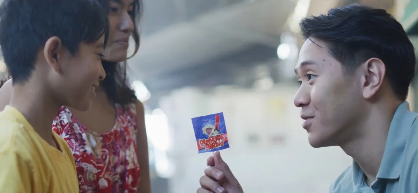 A young man is offering a small packet of snack to two children, a girl and a boy, who are smiling and looking at the snack. The scene is outdoors with a blurred background.