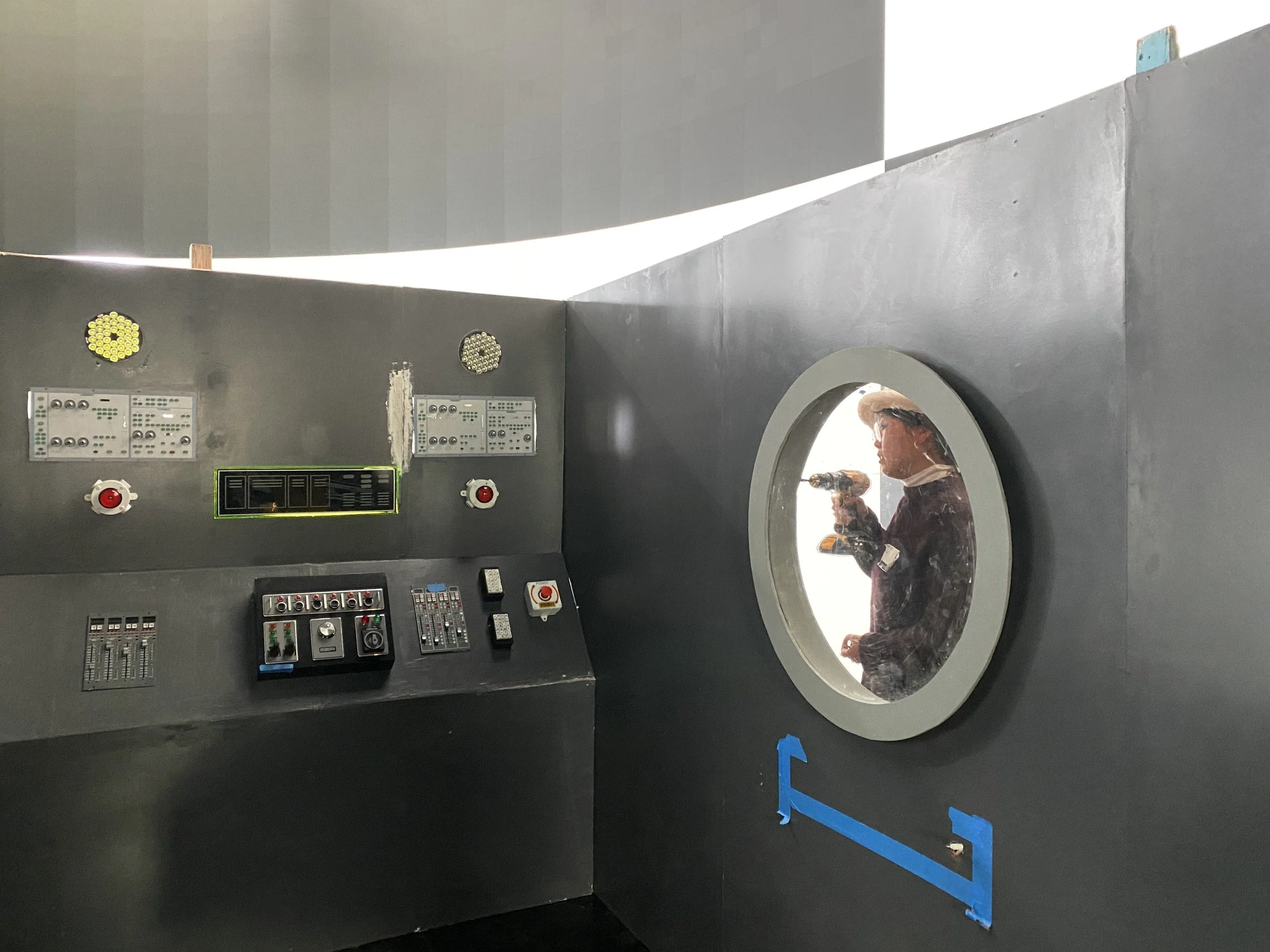A person wearing a protective suit and hat is seen through a circular window, holding a power drill. The interior includes panels with switches, dials, and buttons, resembling a control room or testing chamber.