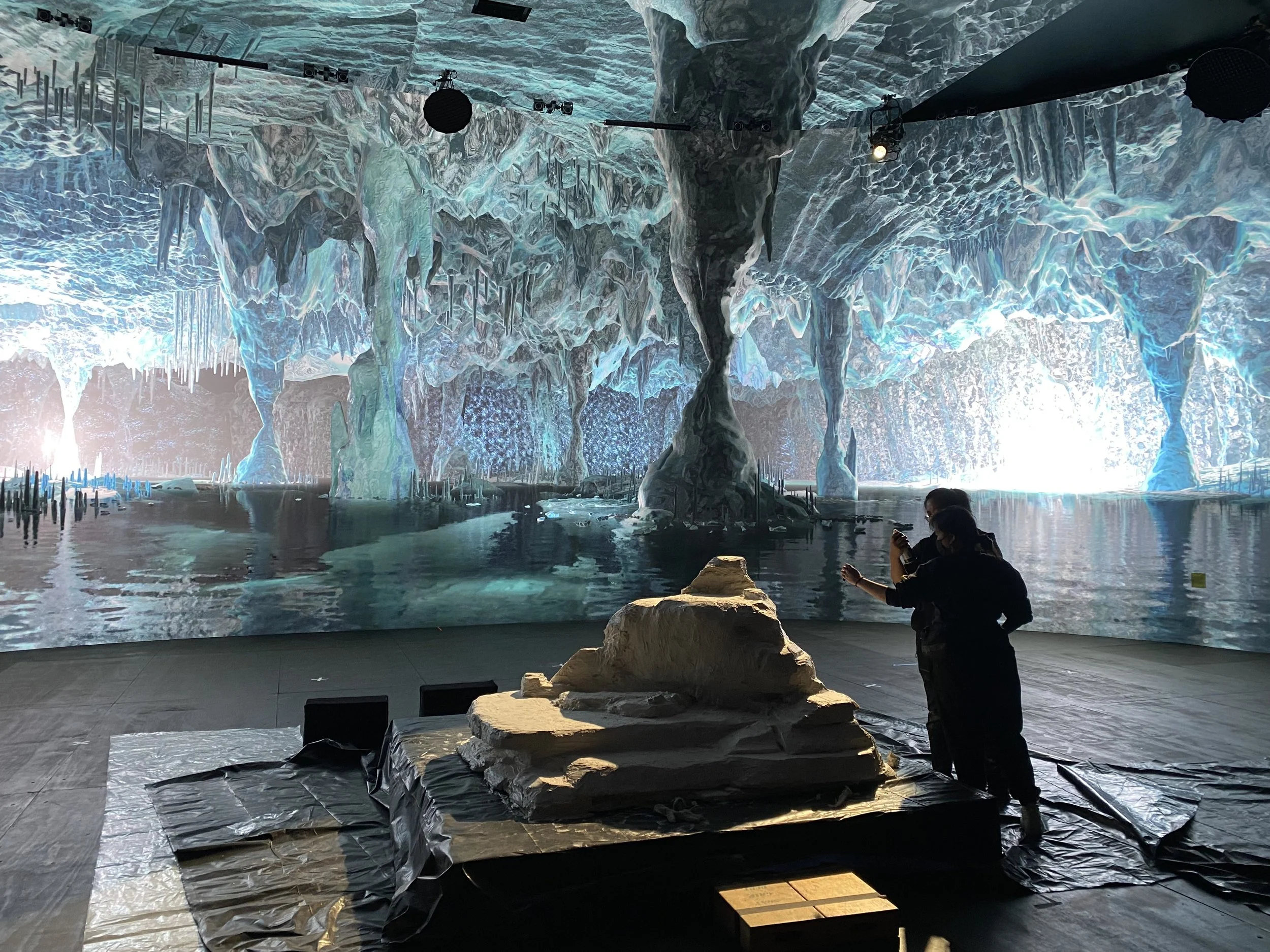 People taking pictures of a large, illuminated virtual ice cave with glaciers and stalactites, with a rock and a small glass display in the foreground.