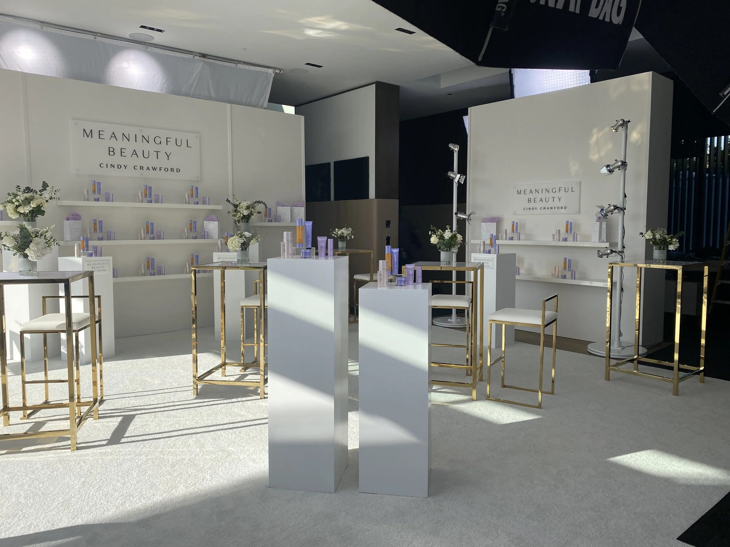 A display booth for 'Meaningful Beauty' by Cindy Crawford, with shelves holding beauty products, floral arrangements, and seating with white cushions and gold frames, set up indoors with professional lighting equipment.