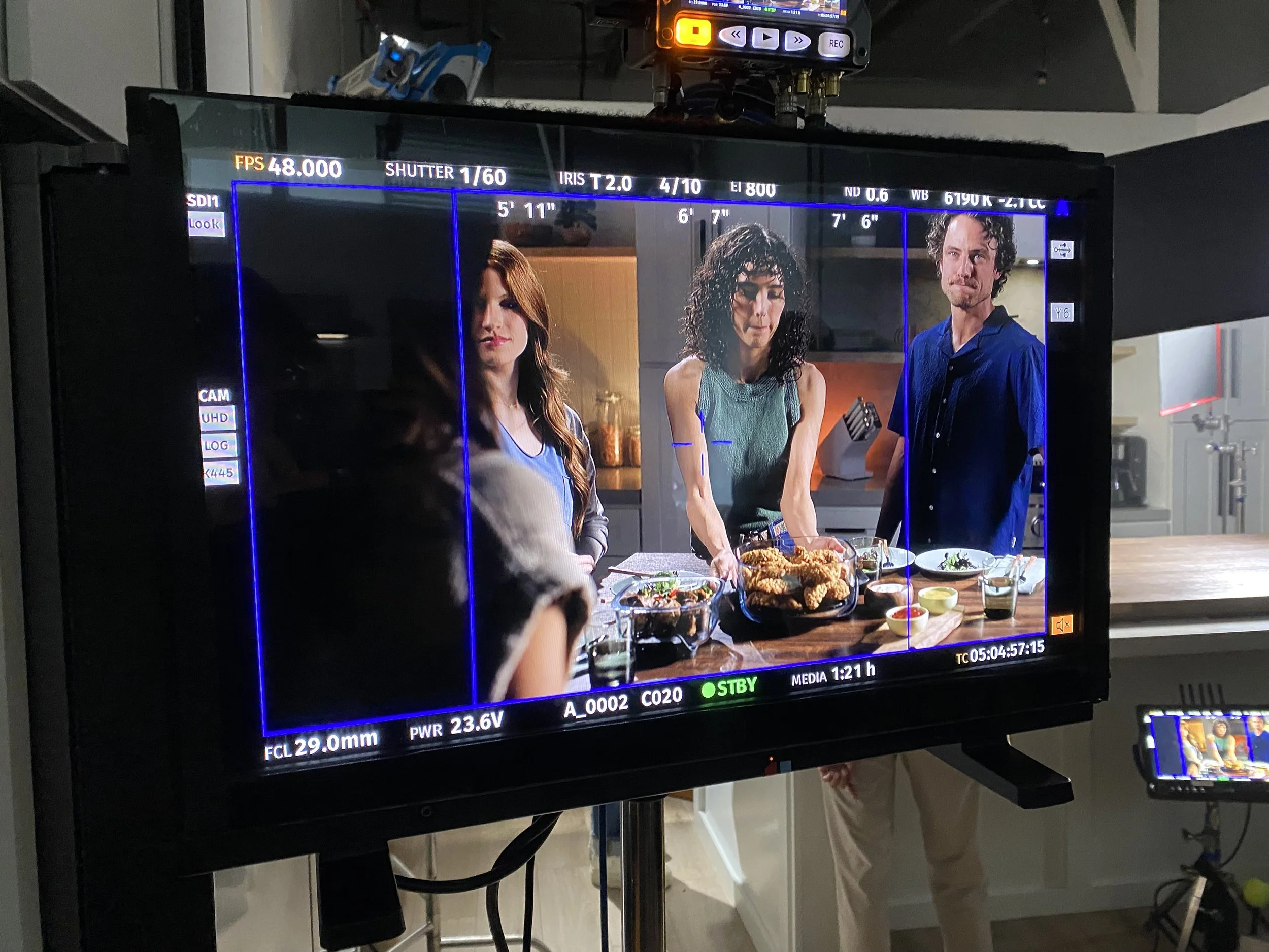 A scene from a film or TV show being filmed, showing three actors around a kitchen counter with plates of food, seen through a monitor screen on a film set.