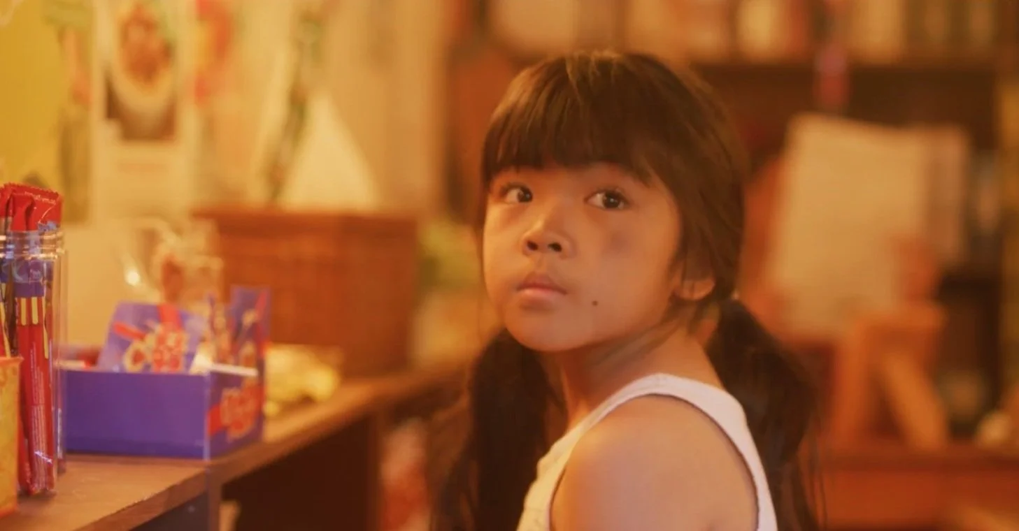 A young girl with long dark hair and bangs sitting in a store with shelves of candy and snacks behind her, looking directly at the camera.