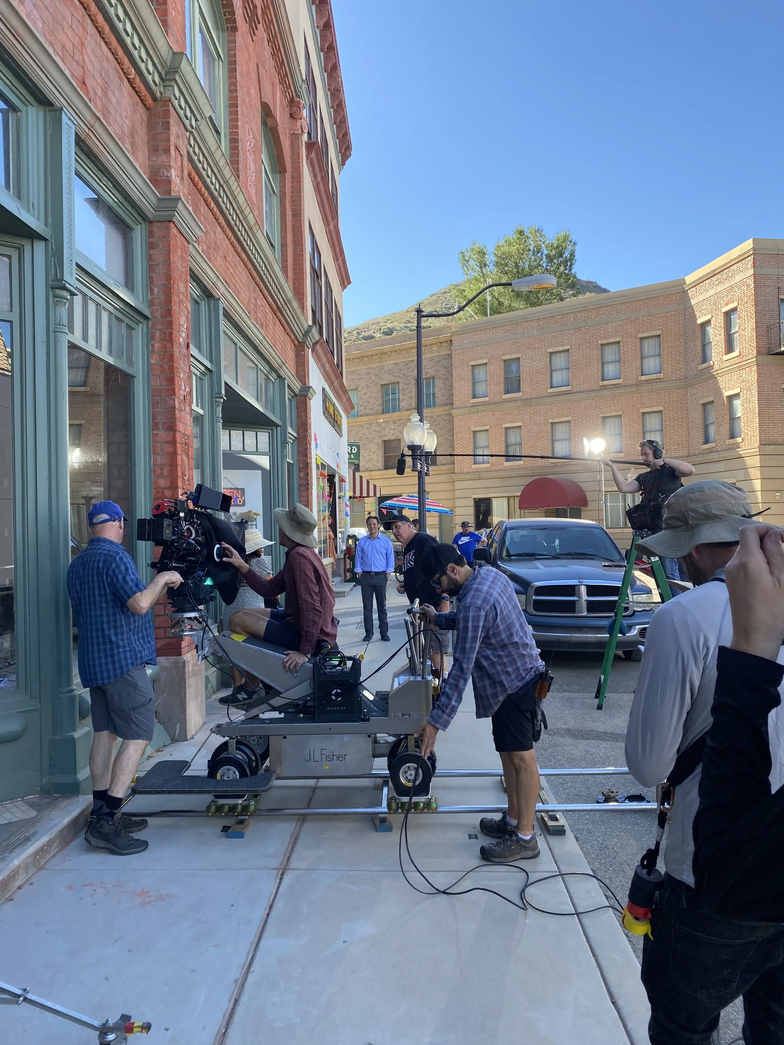 Film crew setting up a shot on a city sidewalk, with a person sitting on a dolly, surrounding crew members adjusting equipment, a camera operator filming, and buildings in the background.