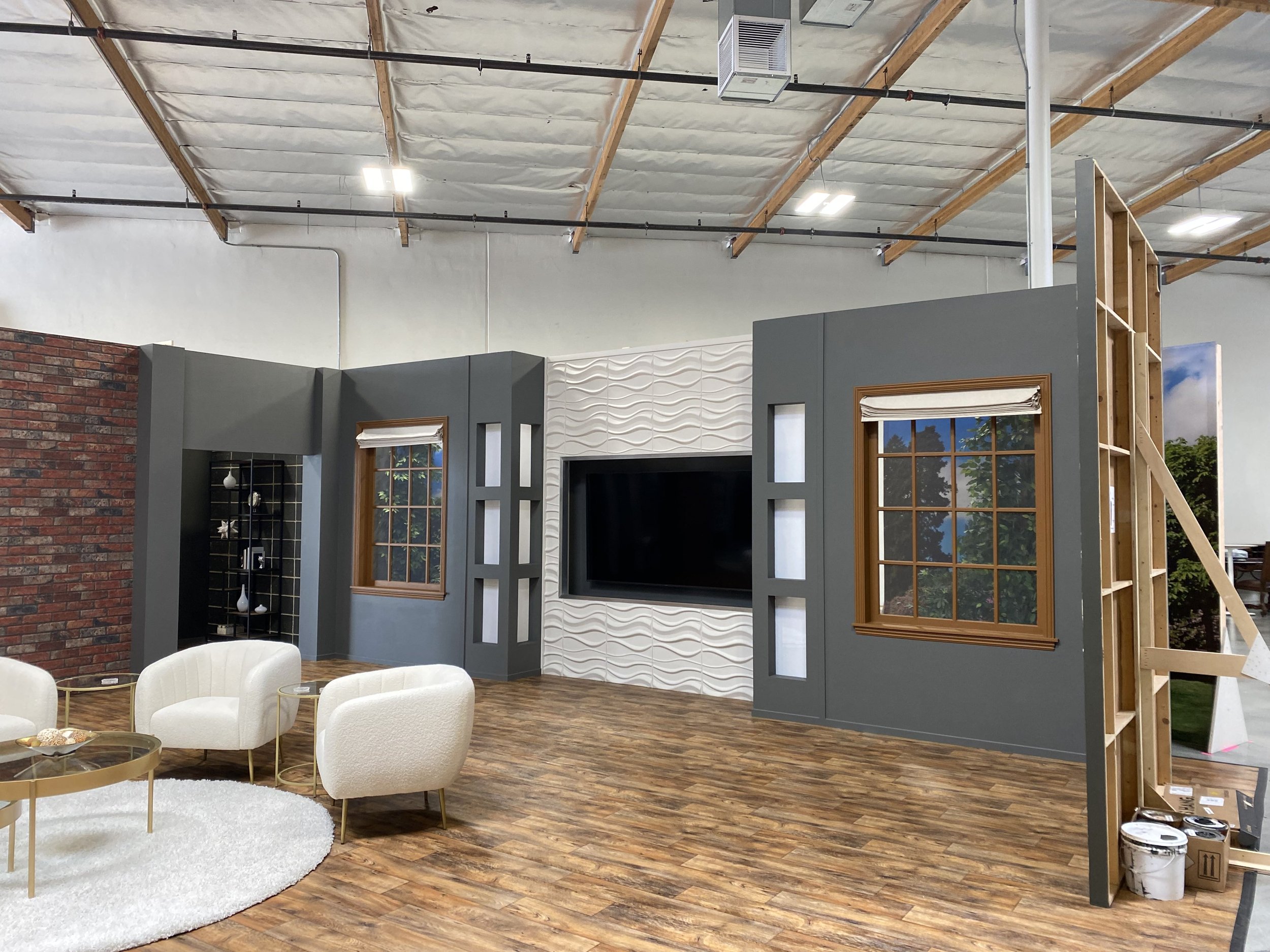 Indoor space resembling a living room with white armchairs, a round white rug, coffee table, and a partial set of walls with windows and decorative panels. The room appears to be under construction or renovation.