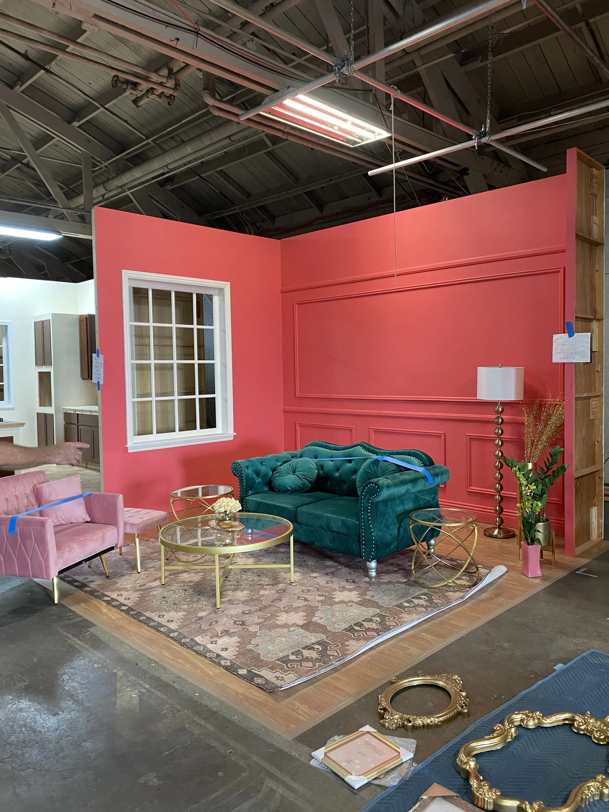Indoor furniture display with pink and green sofas, gold and glass coffee tables, a patterned rug, a tall gold floor lamp, and a decorative potted plant, with a bright pink wall in the background.