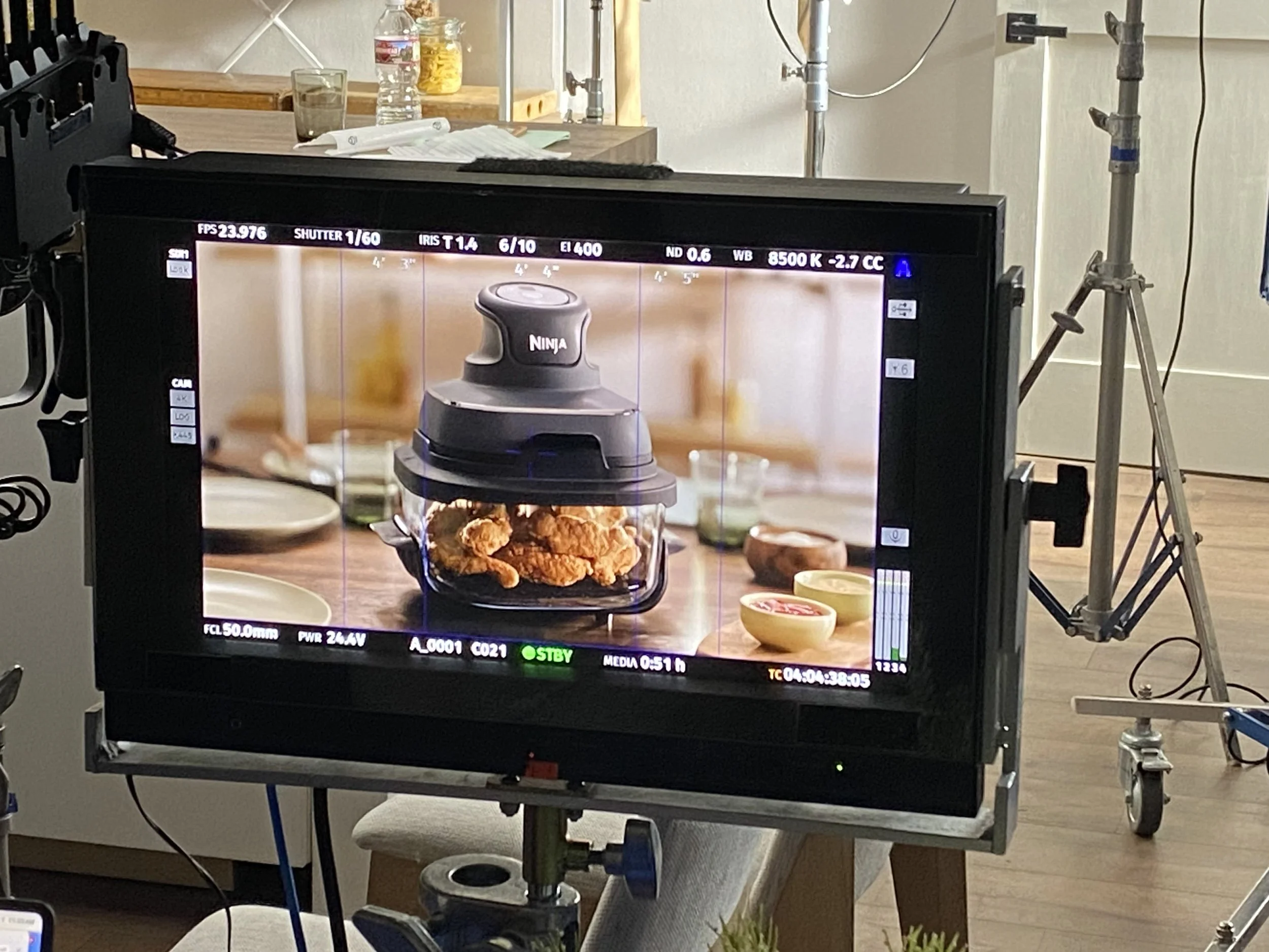 A camera monitor showing a shot of fried chicken inside a Ninja food processor, with bowls and ingredients on a wooden table in the background.