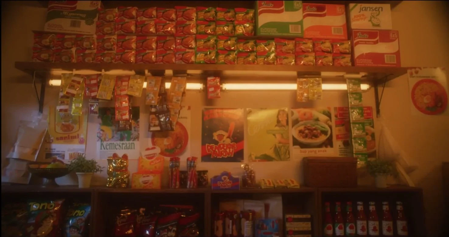 Shelf displaying various snack and condiment packages in a small Asian grocery store, with posters and decorative items on the wall behind.