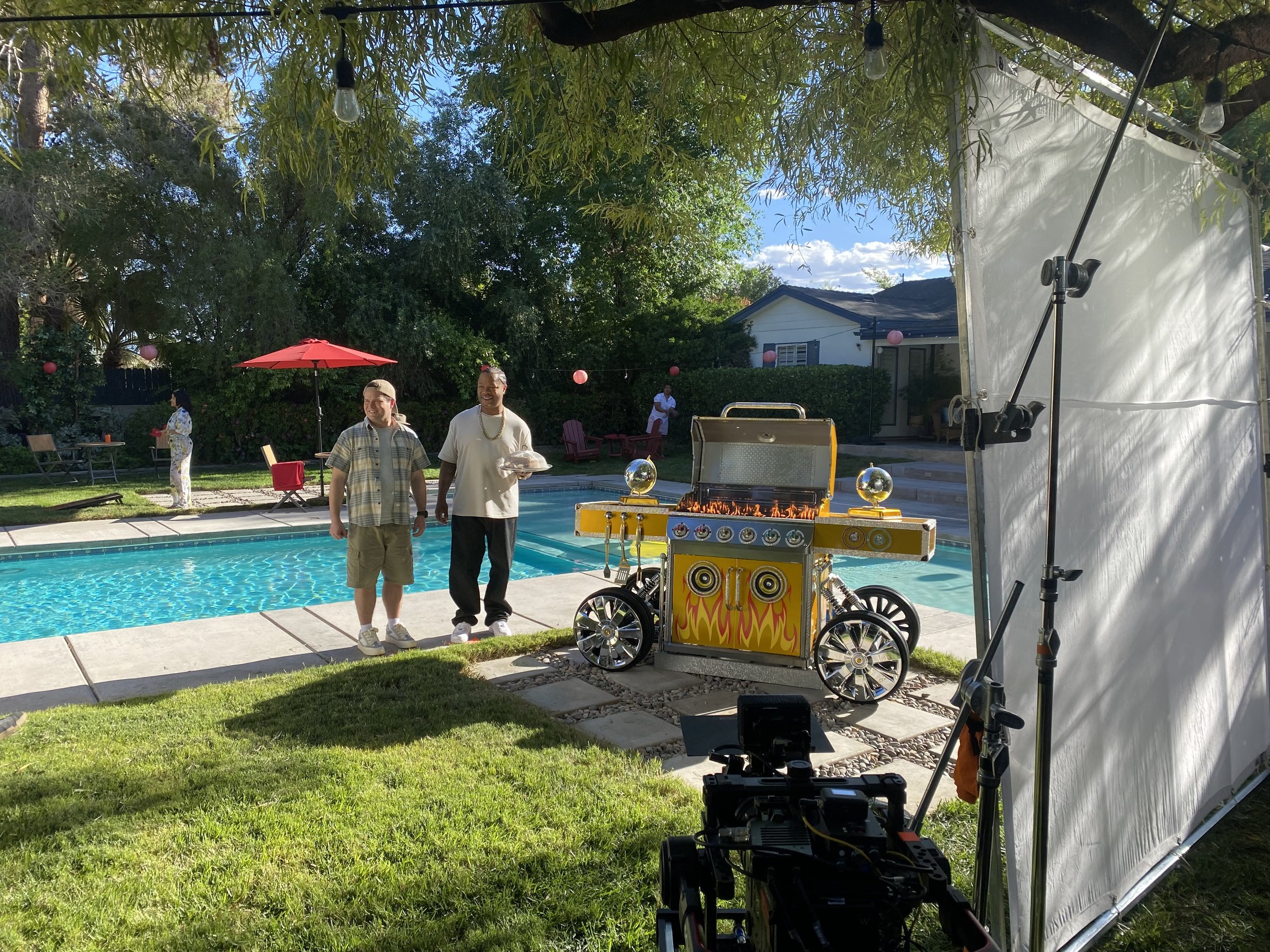 Two men standing by a swimming pool during an outdoor filming session with a portable grill and a backdrop of trees and a house.