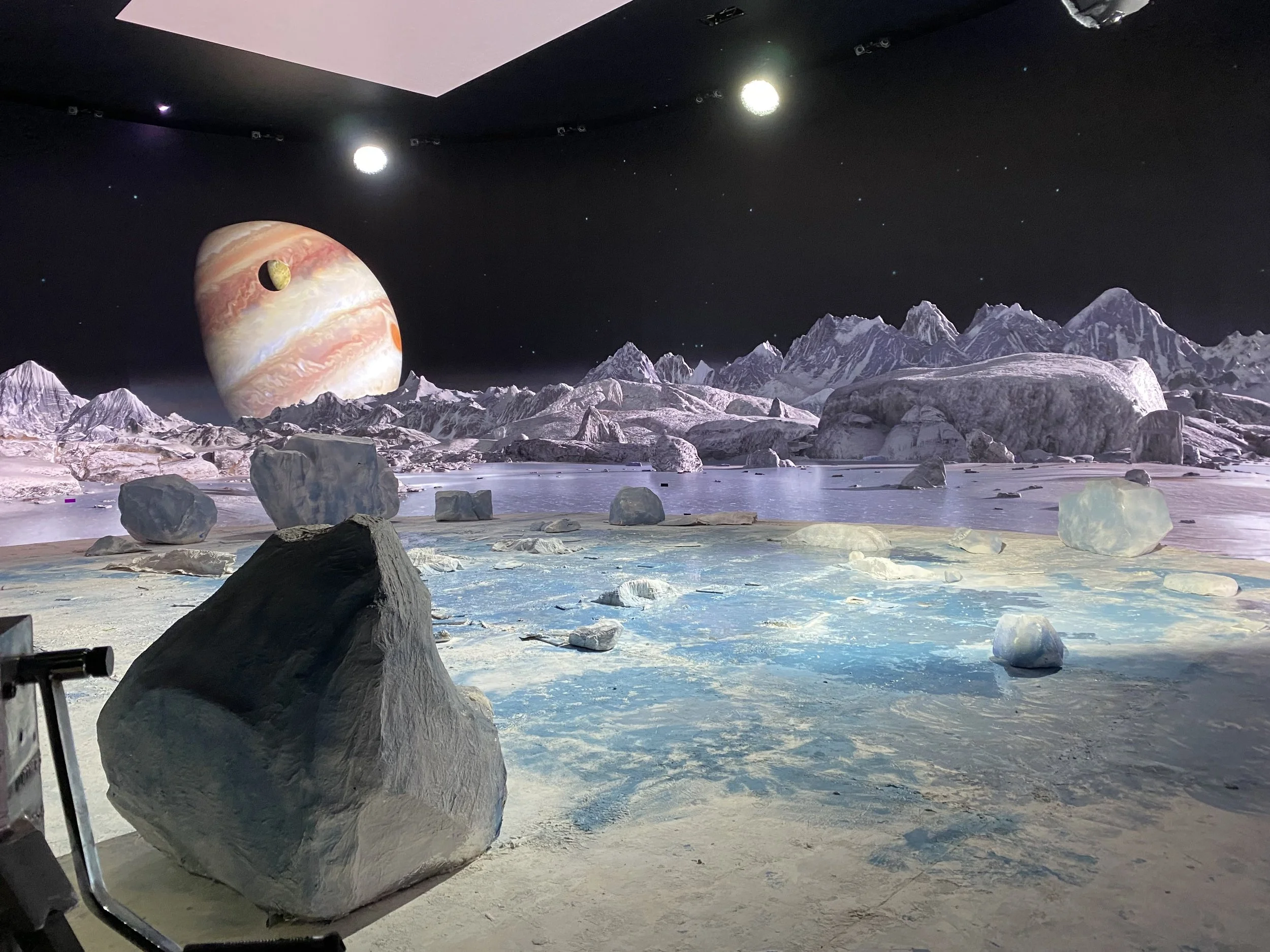 A surreal space scene depicting an icy landscape with large rocks and snow-covered mountains under a starry sky. A giant planet with swirling patterns and a smaller moon are visible in the background.