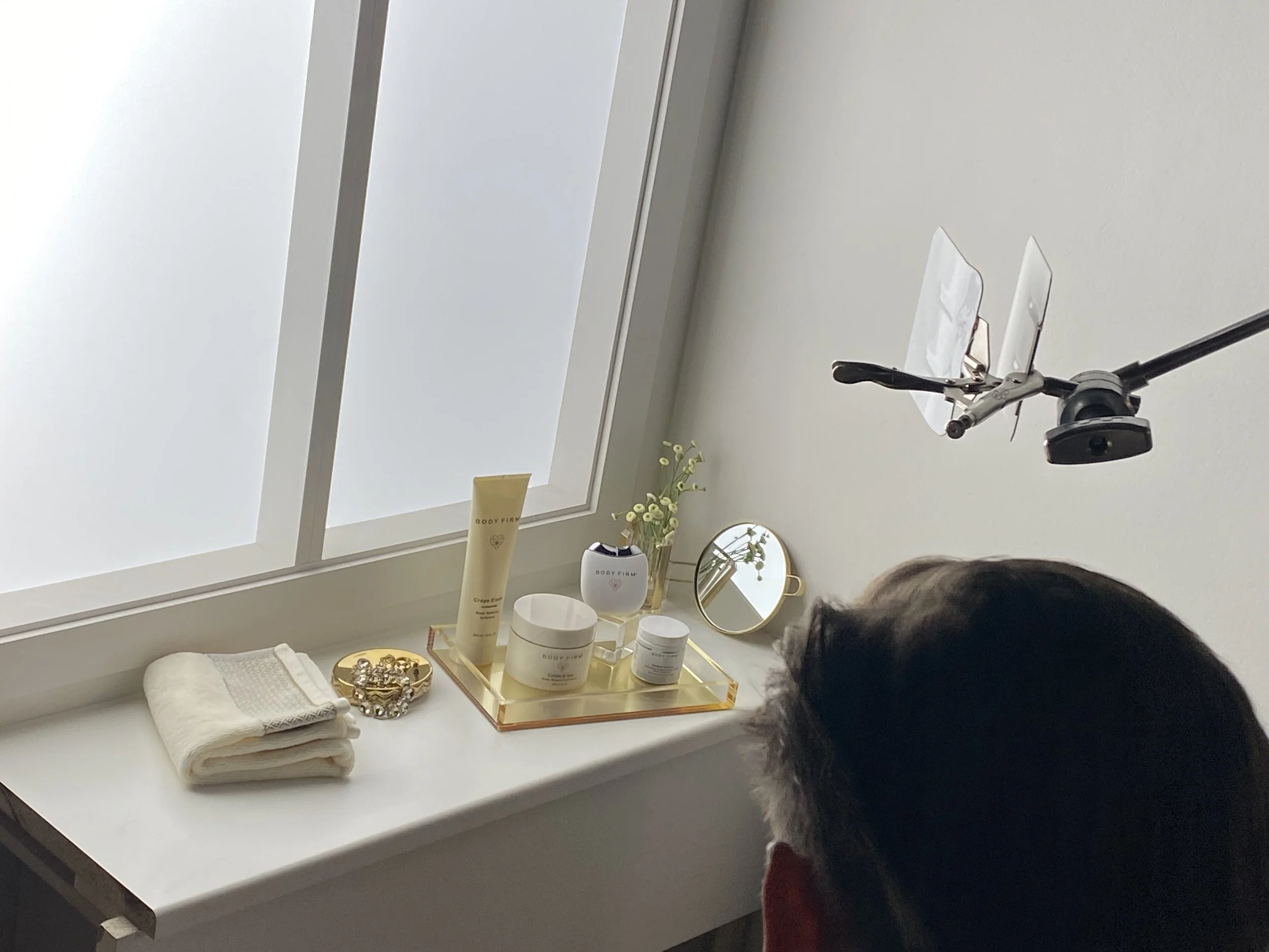 A vanity table by a frosted window with skincare products, a small mirror, a vase with flowers, and a towel. A person with dark hair is seated in front of it, and a camera is mounted above, recording or taking photos.