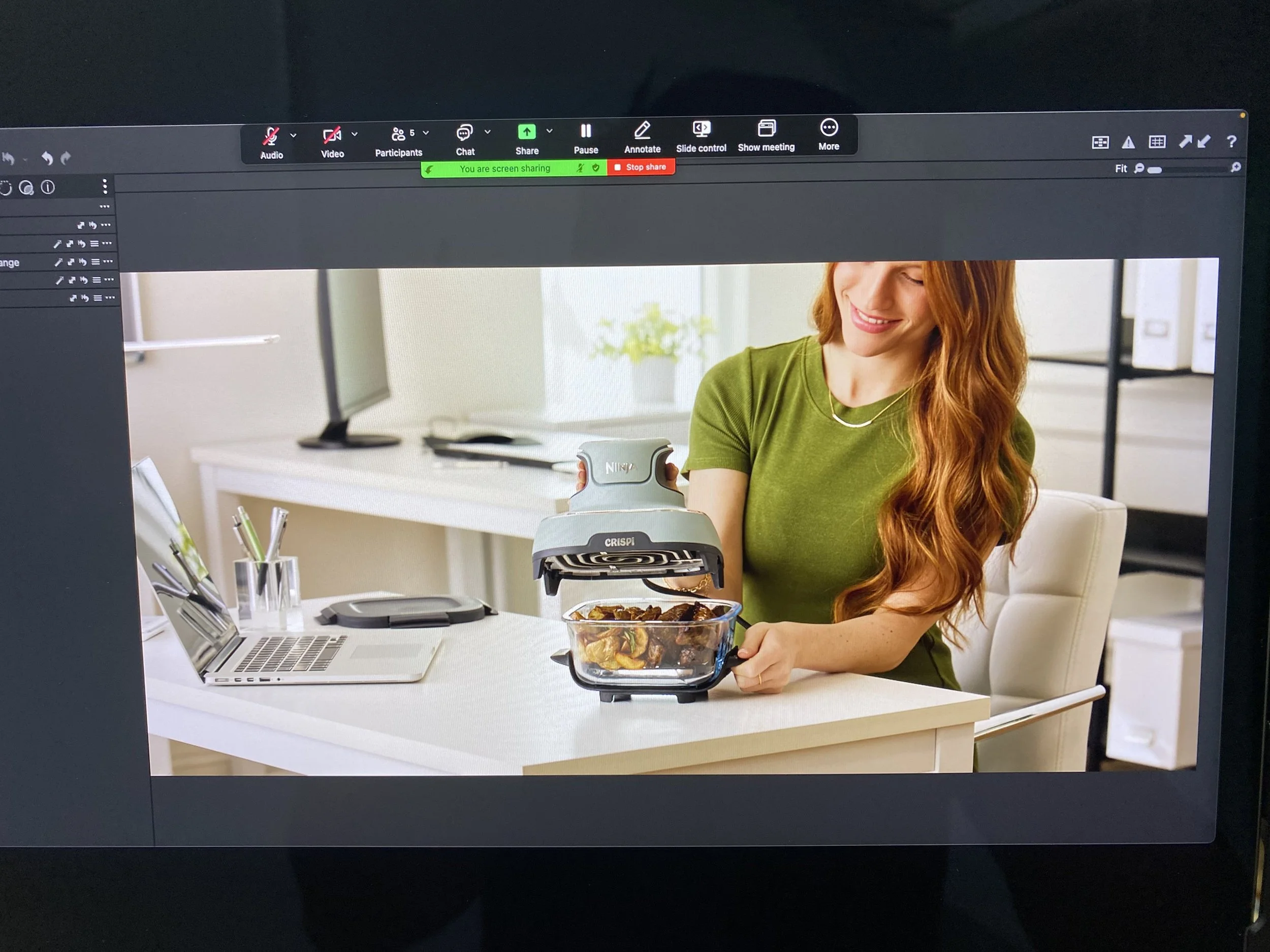 A woman with long red hair wearing a green shirt is using a handheld food steamer to warm up food in a glass container at a white desk, with a laptop, pens, and a monitor in the background.