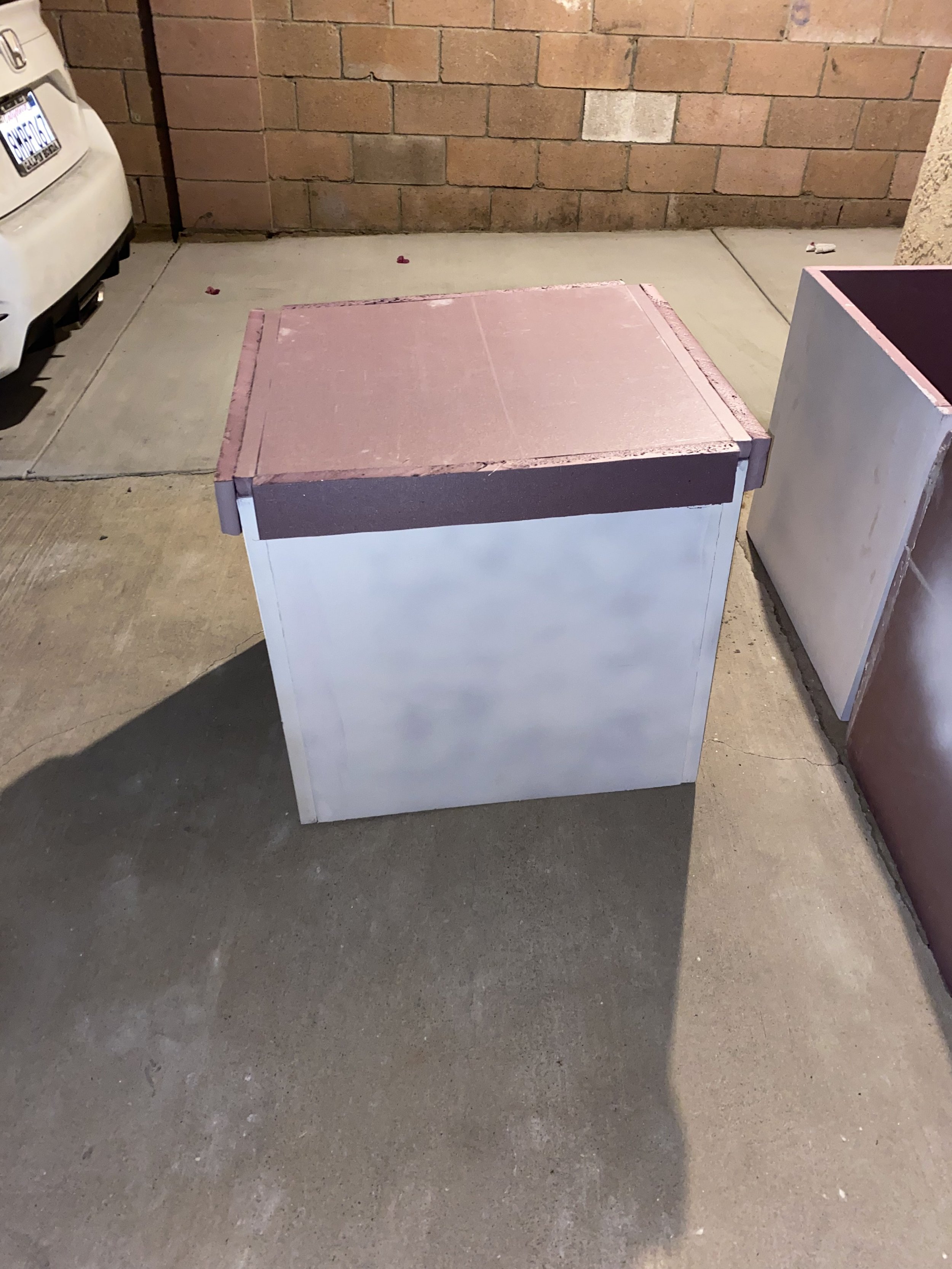 A concrete sidewalk next to a brick wall with a white and pink painted wooden box in the foreground, casting a shadow.