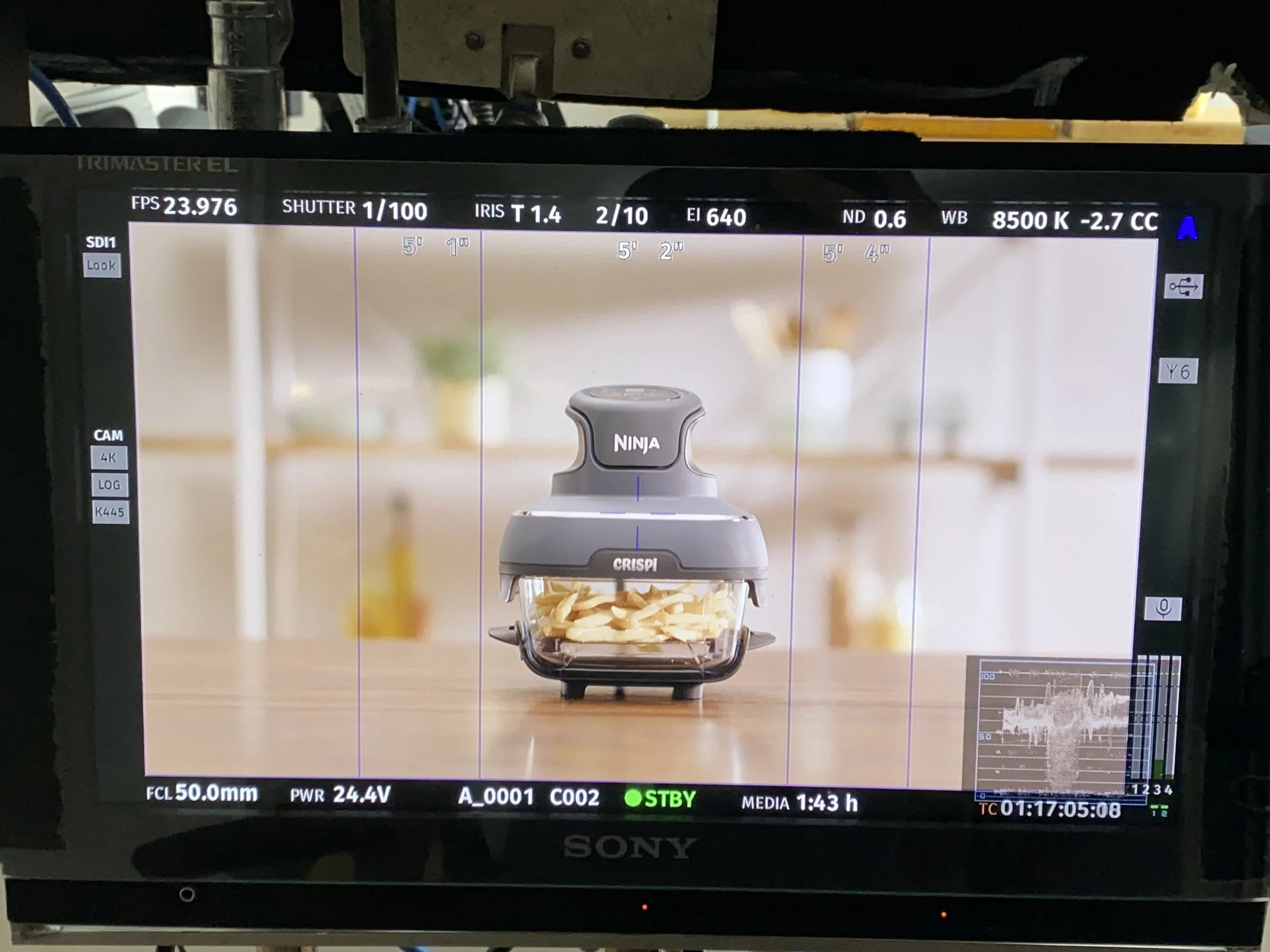 A food processor with the brand name Ninja and the model name Crispi, containing sliced potatoes, is placed on a wooden surface in a kitchen setting.