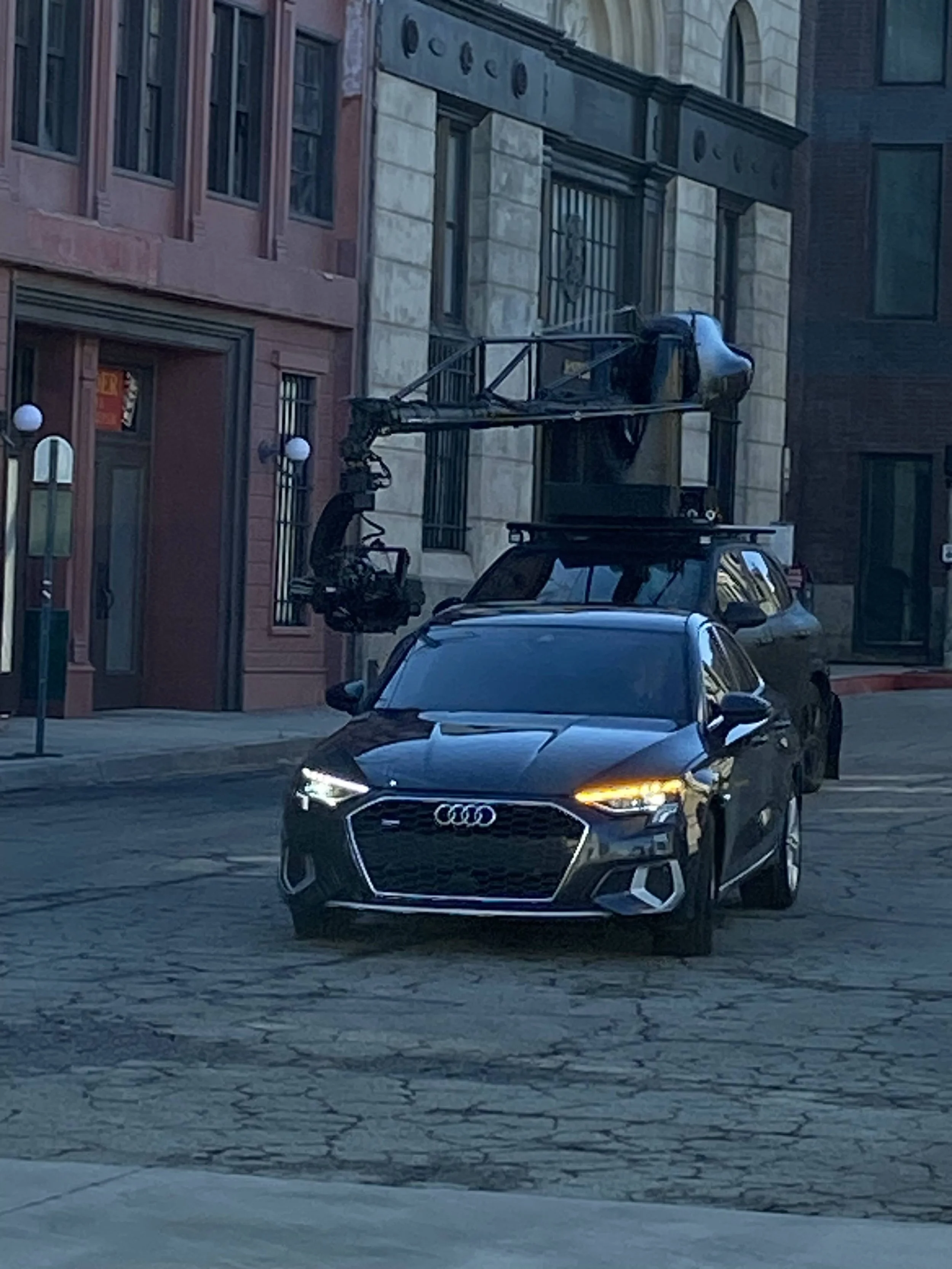 A black Audi car is parked on an urban street, equipped with a mounted camera and crane system on top, possibly used for filming or surveillance, with surrounding buildings and street lamps in the background.
