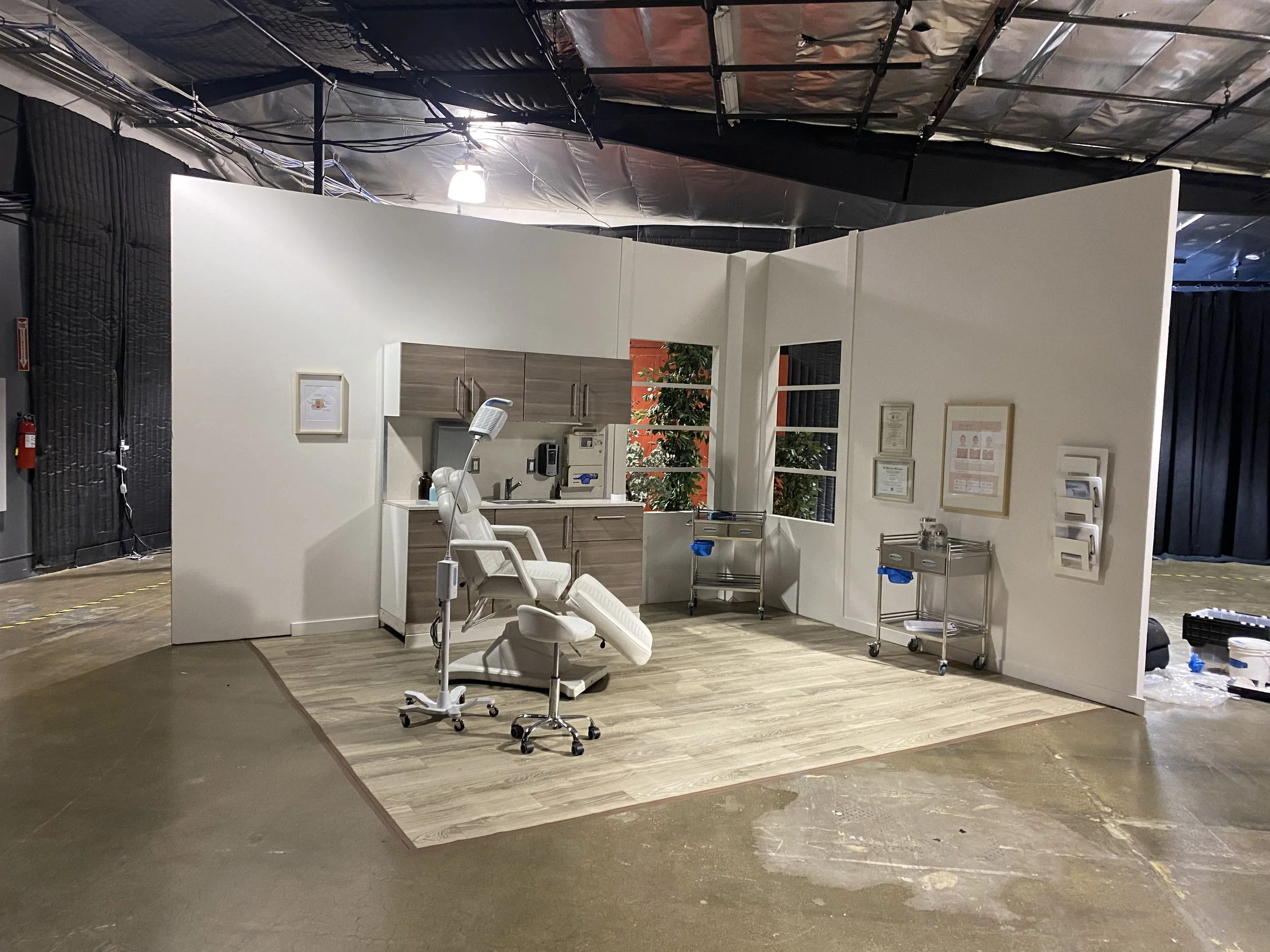 Hospital examination room with a dental chair, medical equipment, and wall-mounted paperwork, set up on a wooden floor with a white partition background.