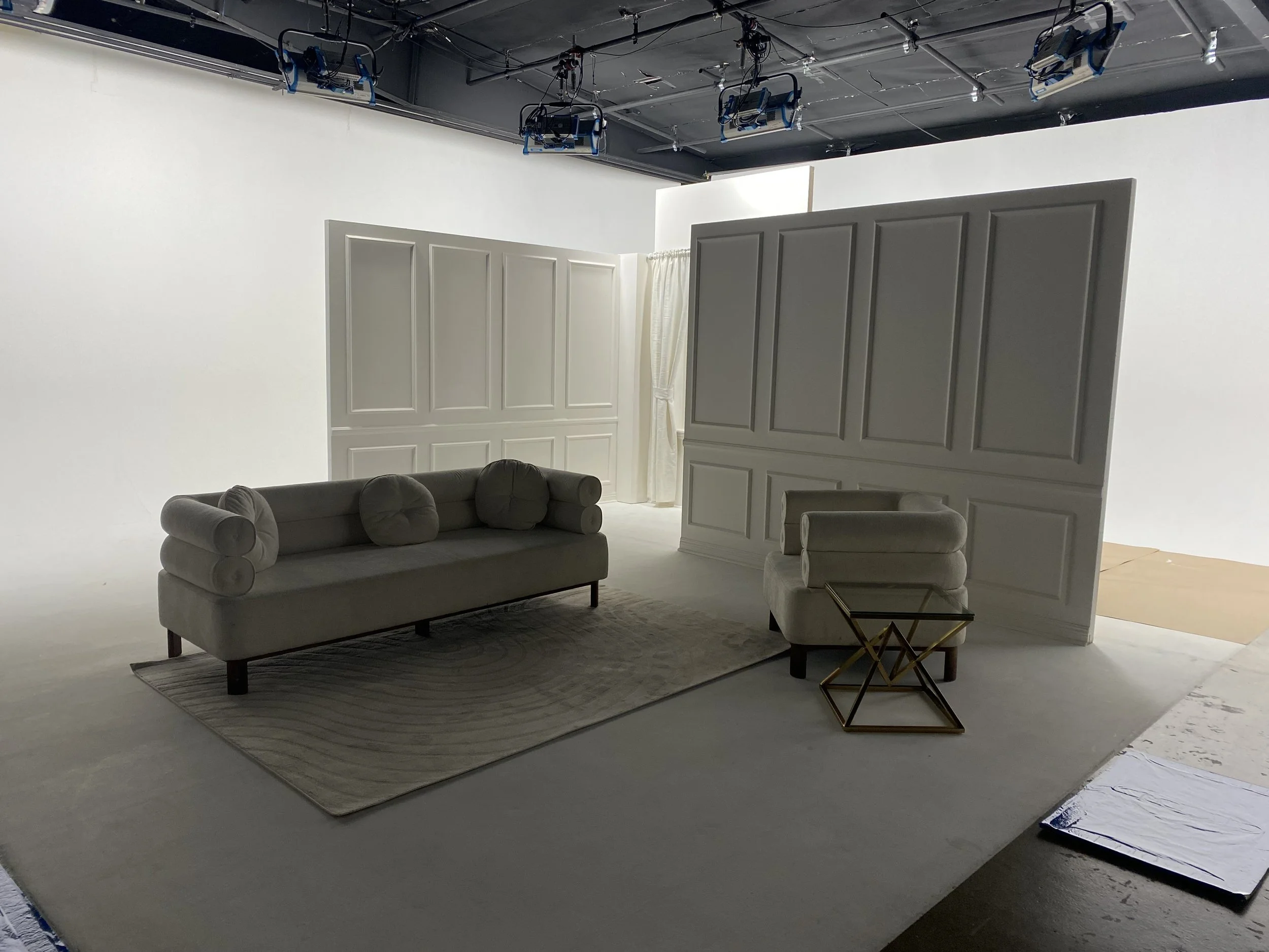 A white television or photography studio set with a beige sofa with round armrests and three cushions, a matching armchair, a small glass-top side table with a geometric gold frame, and a patterned area rug on a beige floor. There are white paneled w
