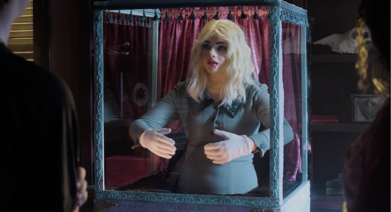 A woman with blonde hair and pale skin, dressed in a gray shirt, inside a glass display case lined with pink velvet curtains, speaking to two people outside the case in a dimly lit room.