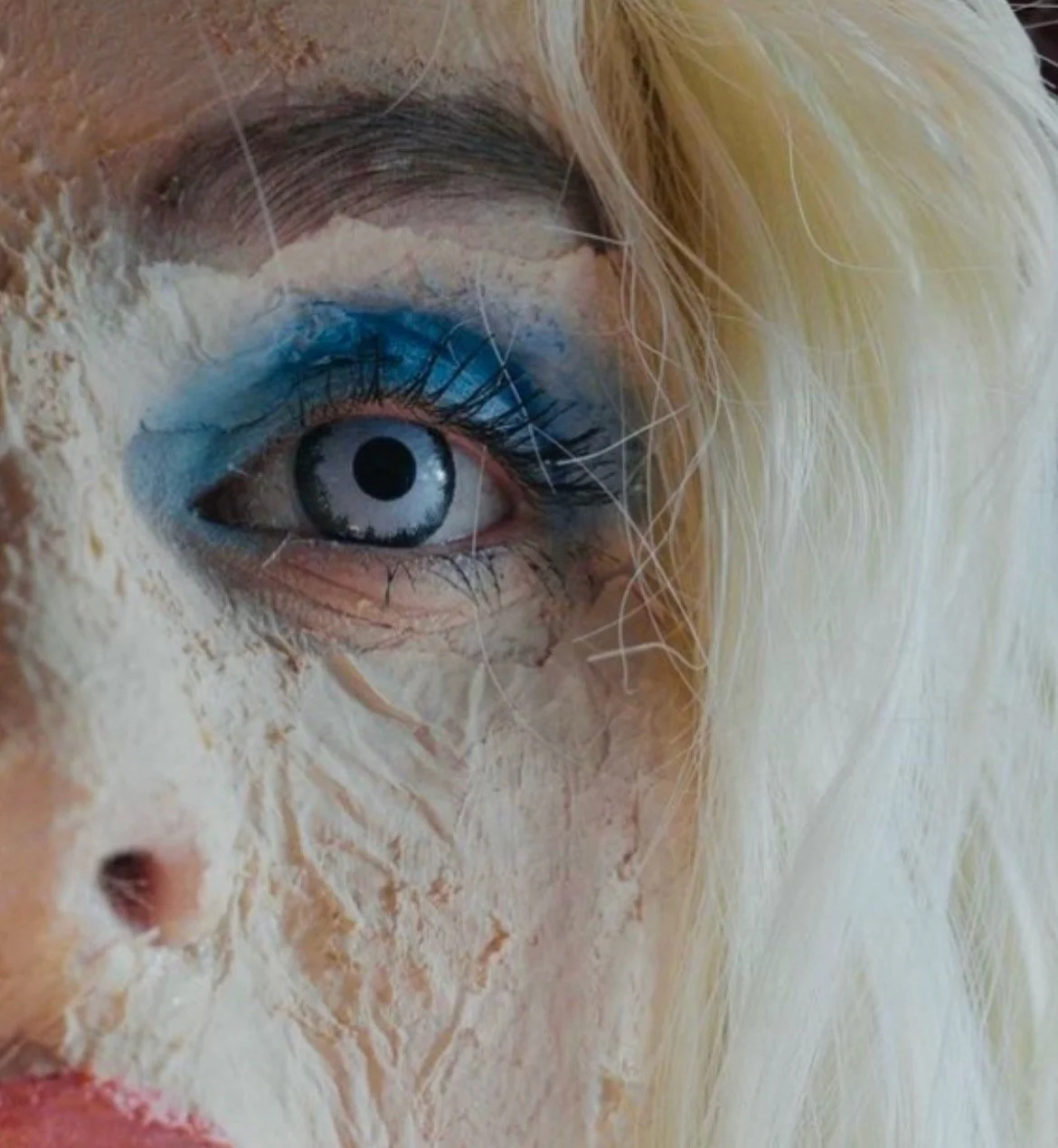 Close-up of a person's eye with blue eyeshadow, surrounded by cracked, peeling white makeup or paint, with blonde hair partially visible.