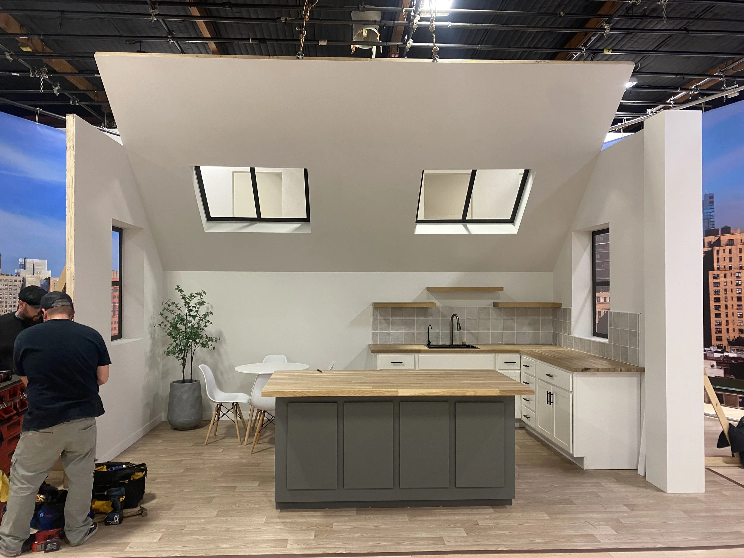 A modern kitchen setup with white cabinets, wooden countertops, and a gray island. Two skylights are on the ceiling, and there is a potted plant and a small dining area with a round table and four chairs. Two people are working on tools on the left s