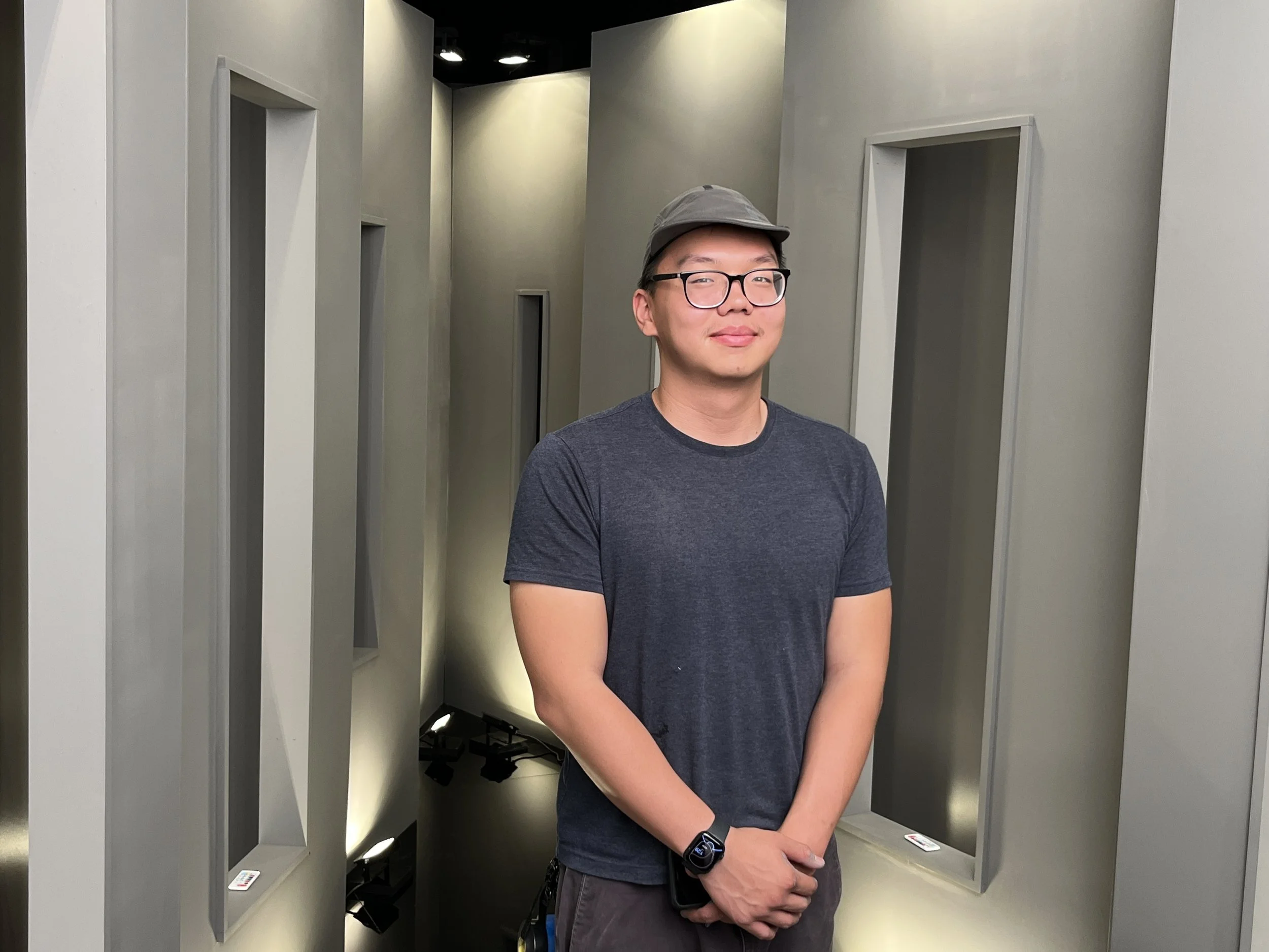 Young man wearing glasses, a gray hat, and a dark gray T-shirt standing in a modern room with gray walls and targeted lighting.