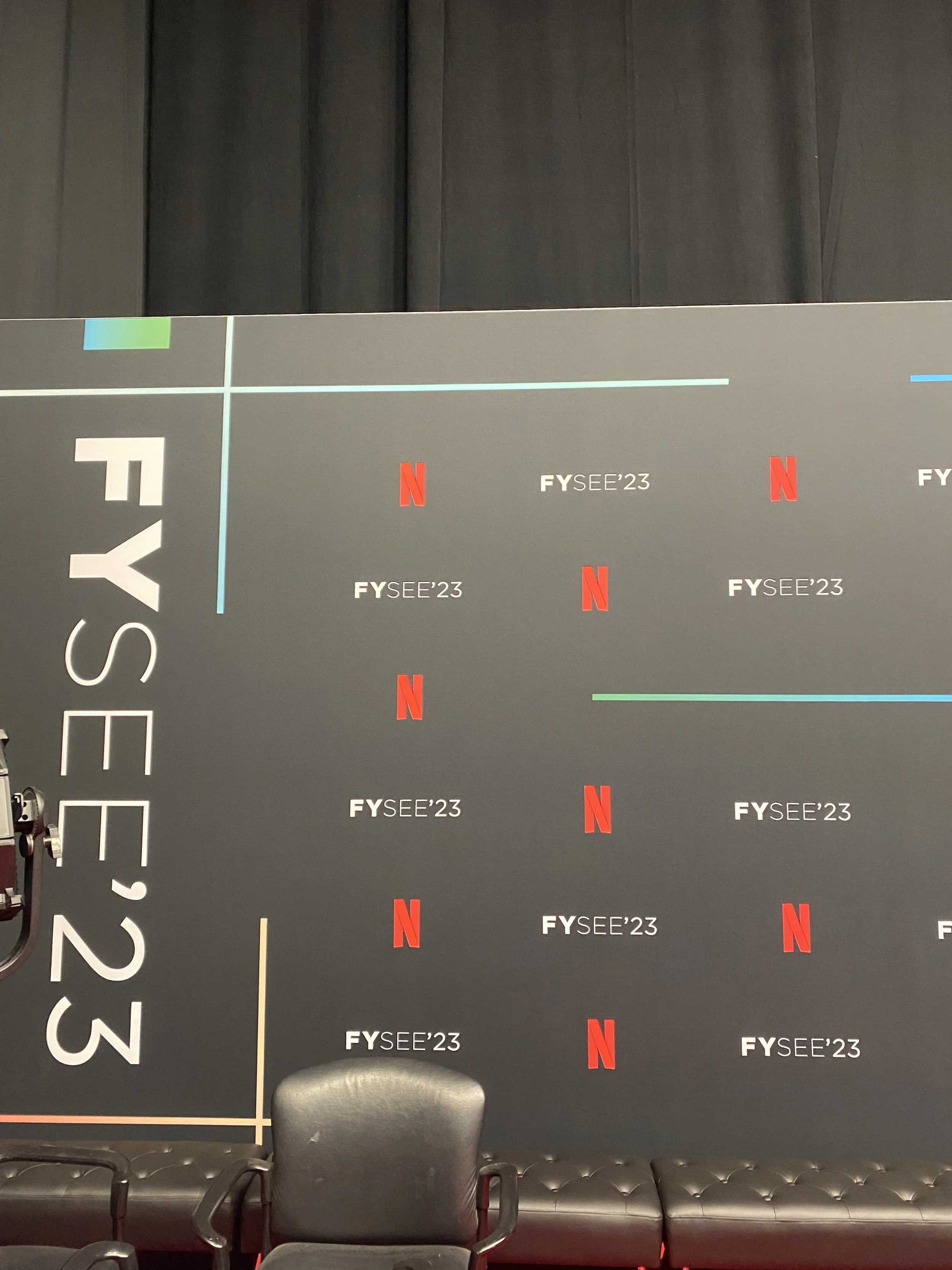 A dark gray wall with the text 'FYSEE' and '23' repeated multiple times, along with red Netflix logos. To the left, there is a large white vertical text that reads 'FYSEE 23'. Part of a black chair and some foam padding are visible at the bottom of t
