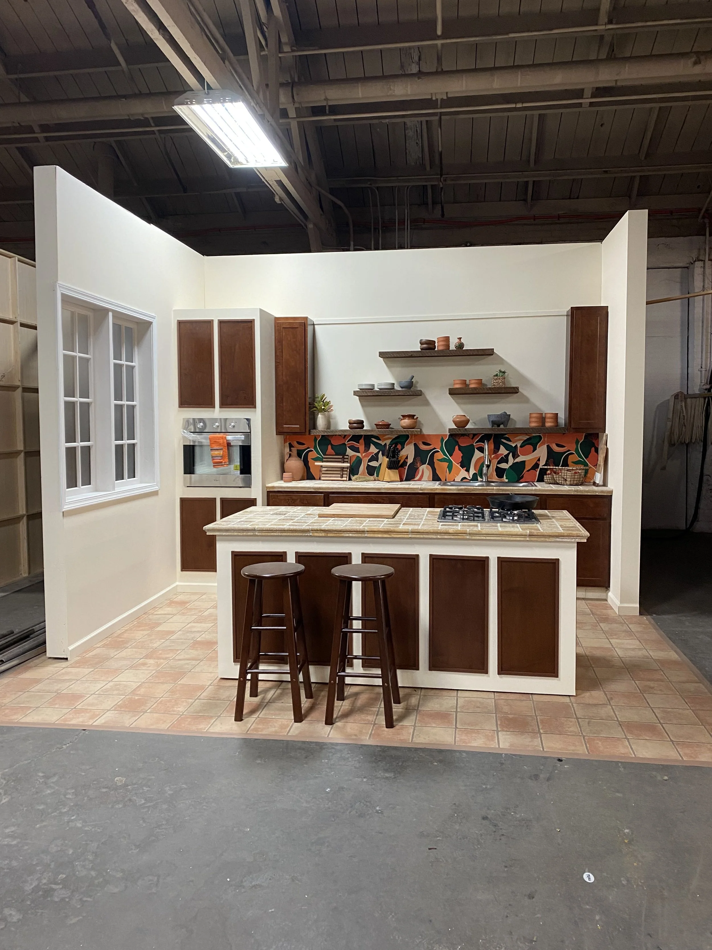 A small kitchen setup display with a tiled island counter and two wooden stools, white walls, dark wooden cabinets, open shelves with pottery, and a decorative backsplash with a colorful pattern.