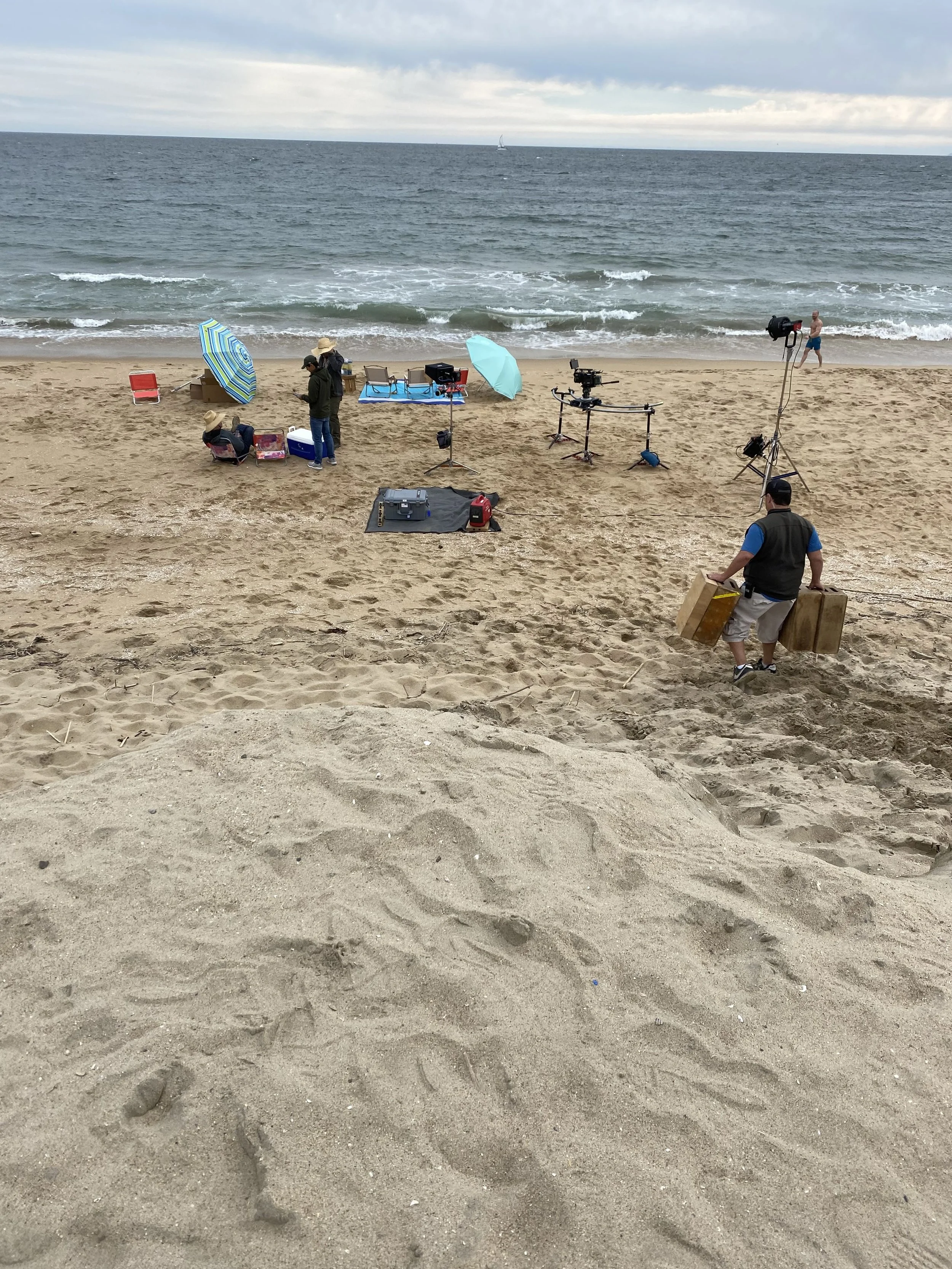 Beach scene with a film crew setting up equipment, some crew members with umbrellas, and a man carrying large boxes walking on the sand towards crew members.