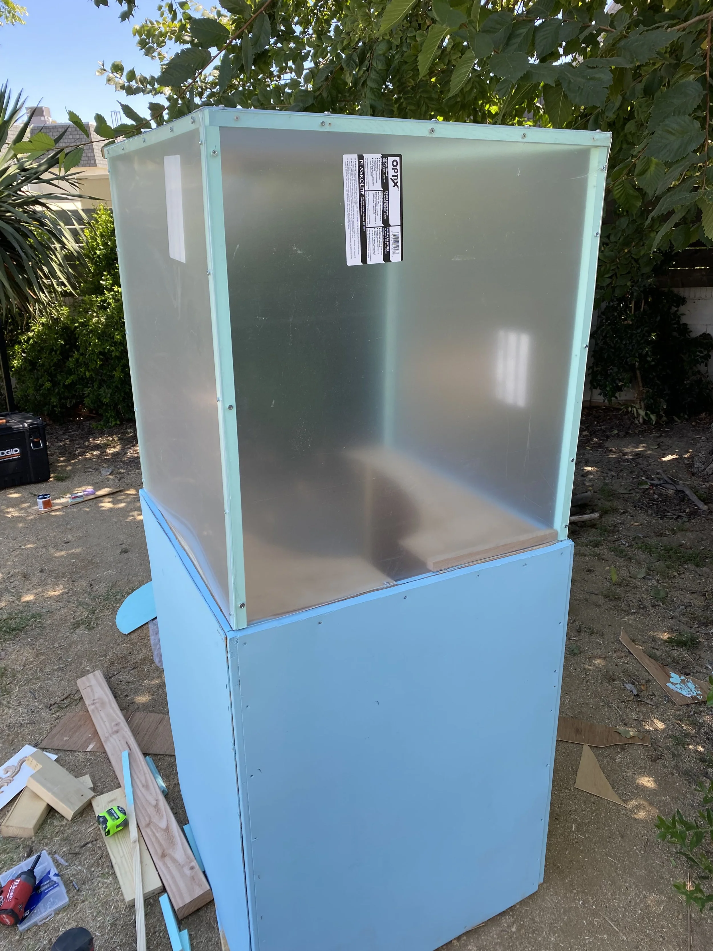 A vintage popcorn machine with a light blue base and a clear glass enclosure on top, situated outdoors in a garden with green foliage.