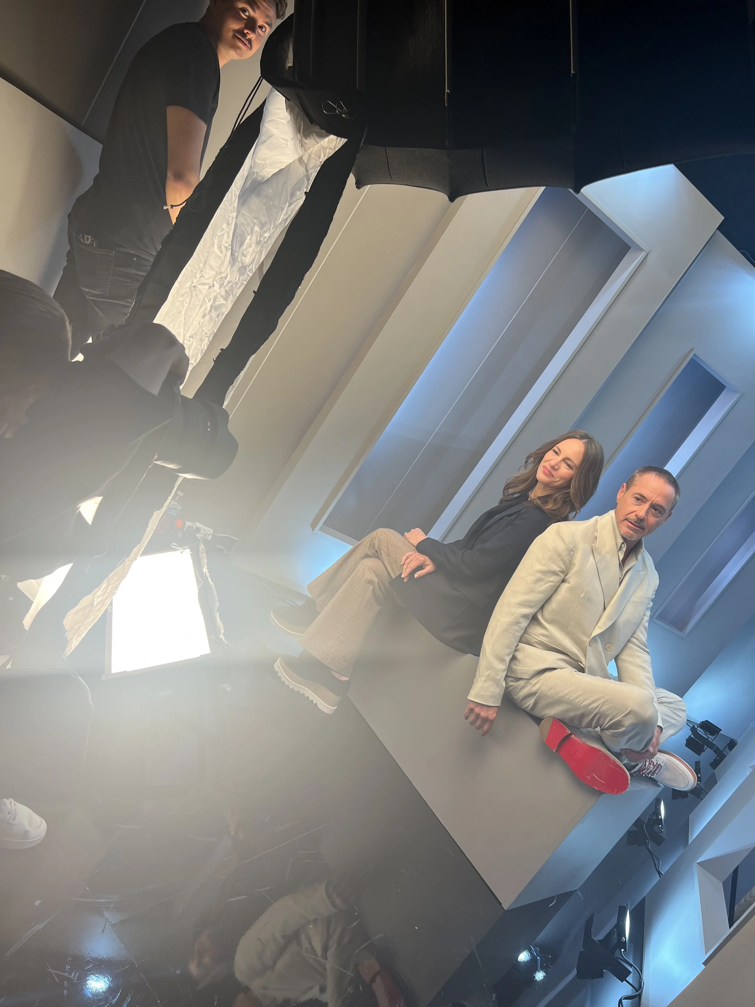 Two people sitting on a platform in a photo studio with lighting equipment and a photographer taking pictures.