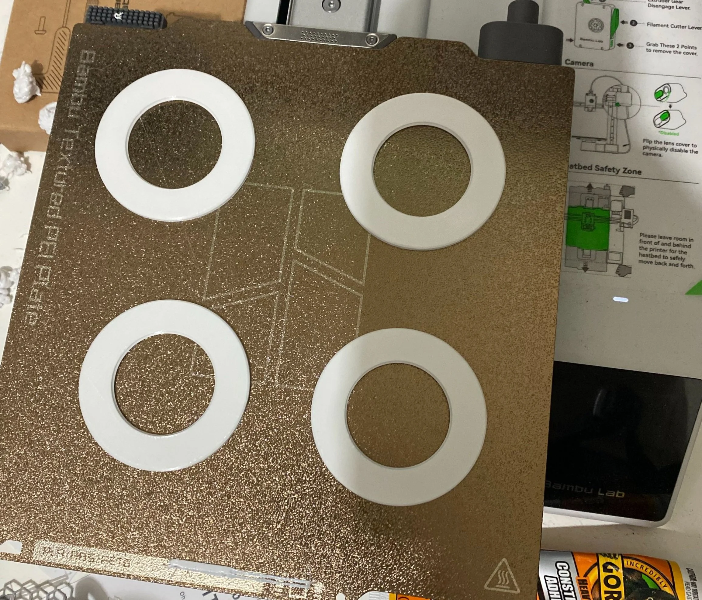 A gold-colored 3D printer build plate with four white washers on top, placed on a cluttered surface with various tools and instruction papers around.