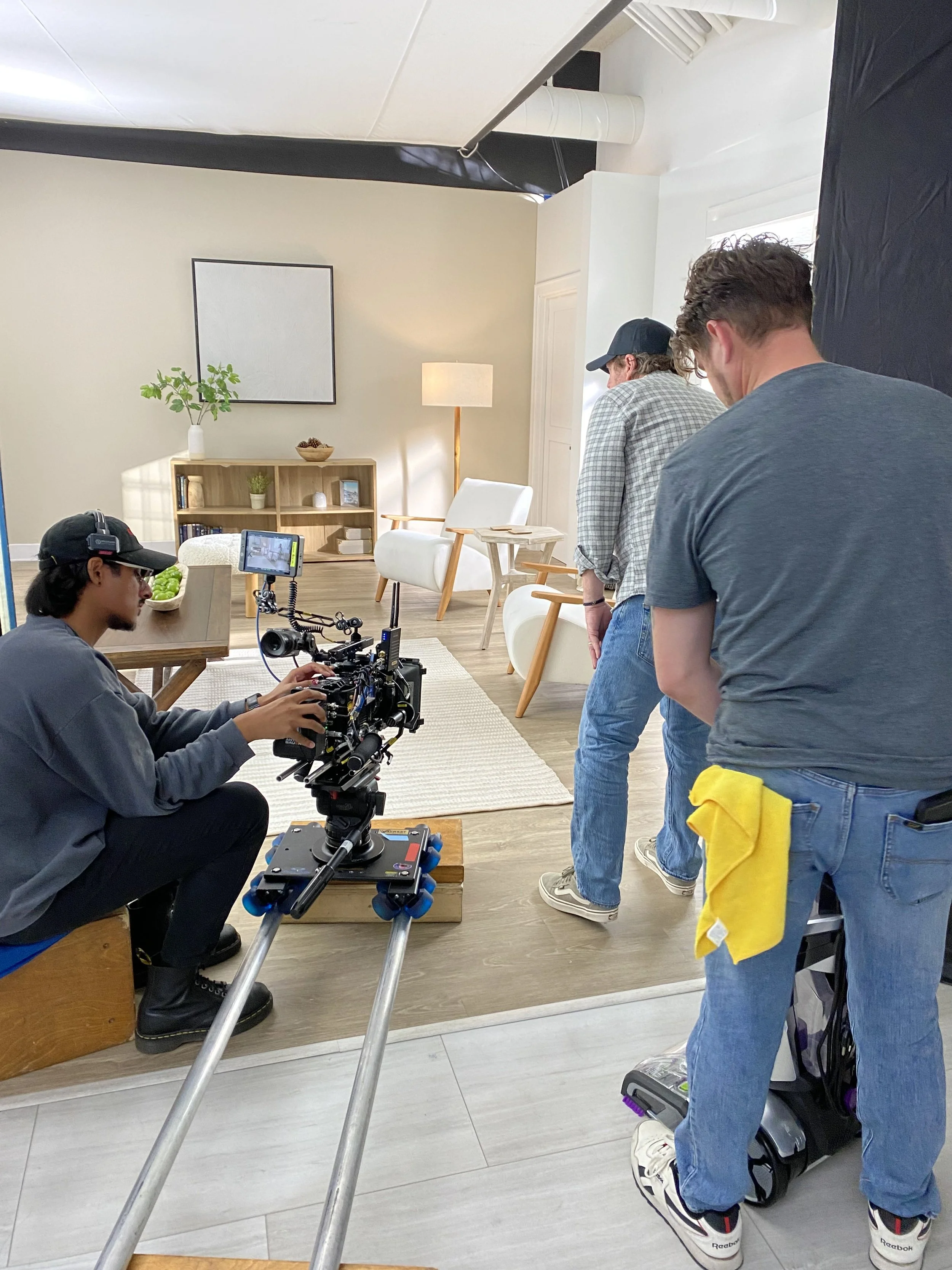 Filming crew setting up in a living room with a camera on a dolly, a person operating the camera, and two other crew members preparing for a shoot.