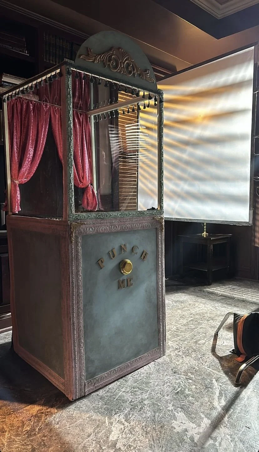 A vintage-style punch and meet booth with red curtains, gold accents, and the words "PUNCH ME" on the front. Sunlight filters through blinds, creating a warm glow.