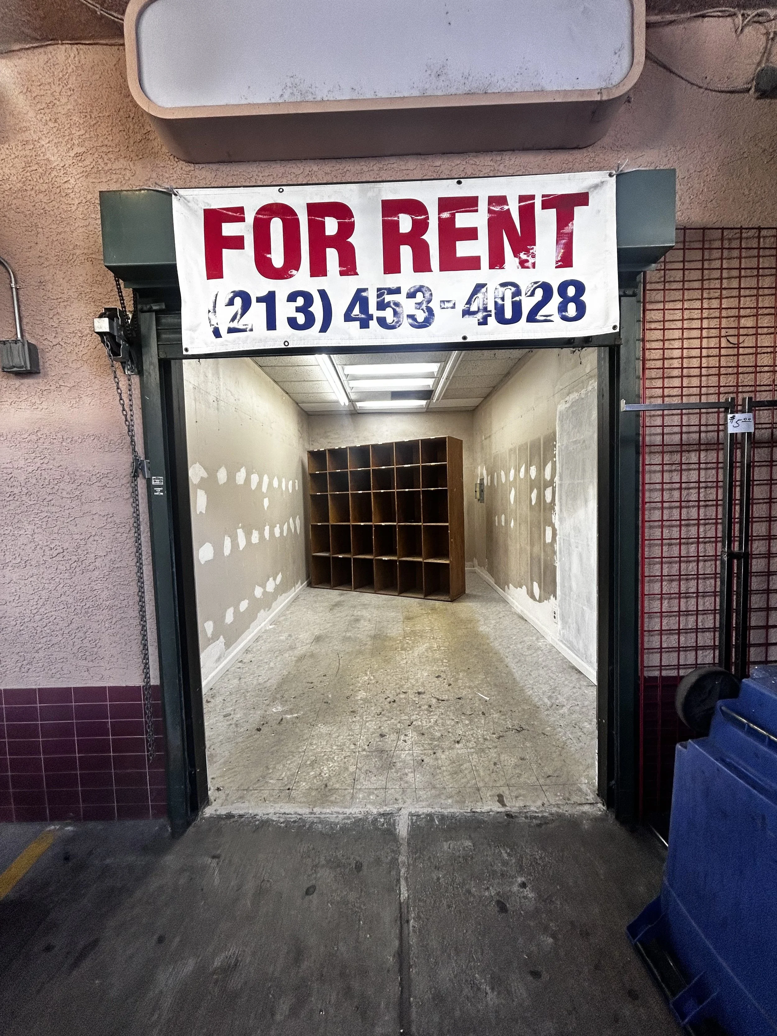 Empty retail space for rent with a banner displaying a phone number, inside the space are wood cubby shelves and unfinished walls.